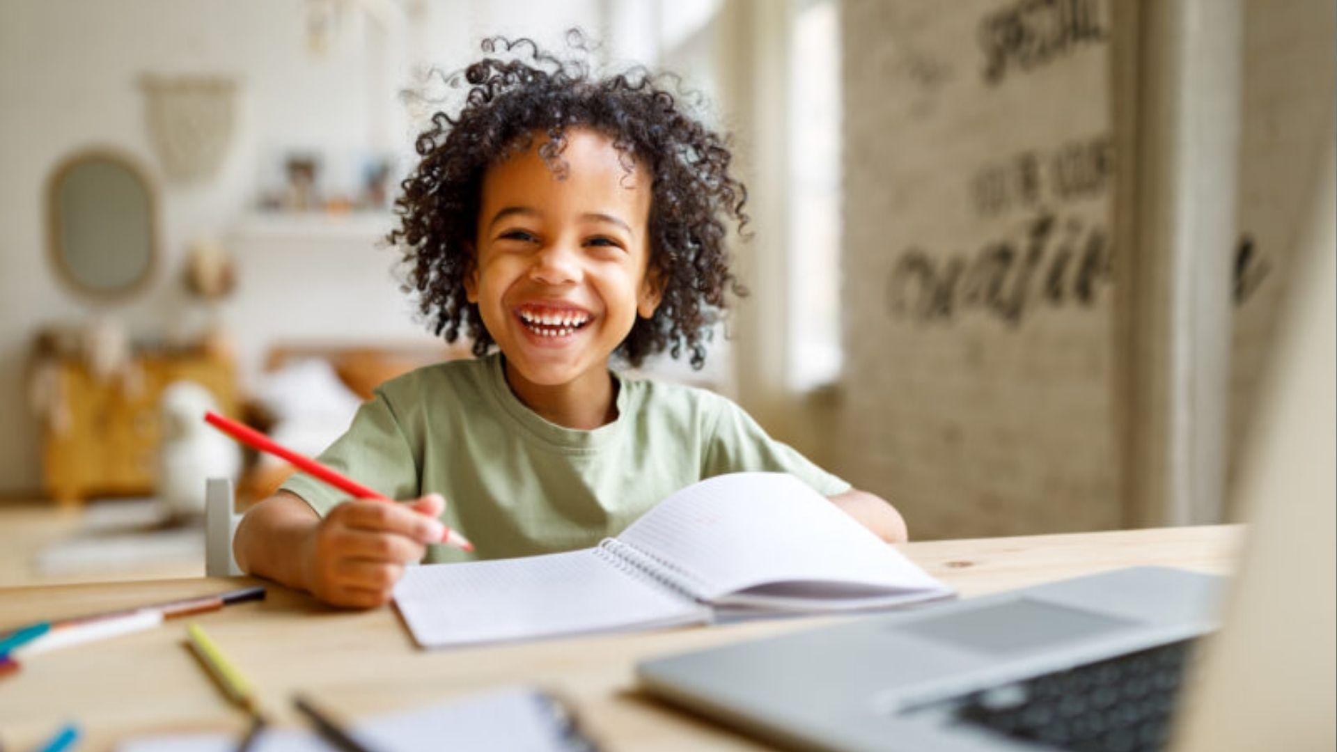 A smiling child holds a red pencil over a notebook, proving fun, relaxed practice improves handwriting.