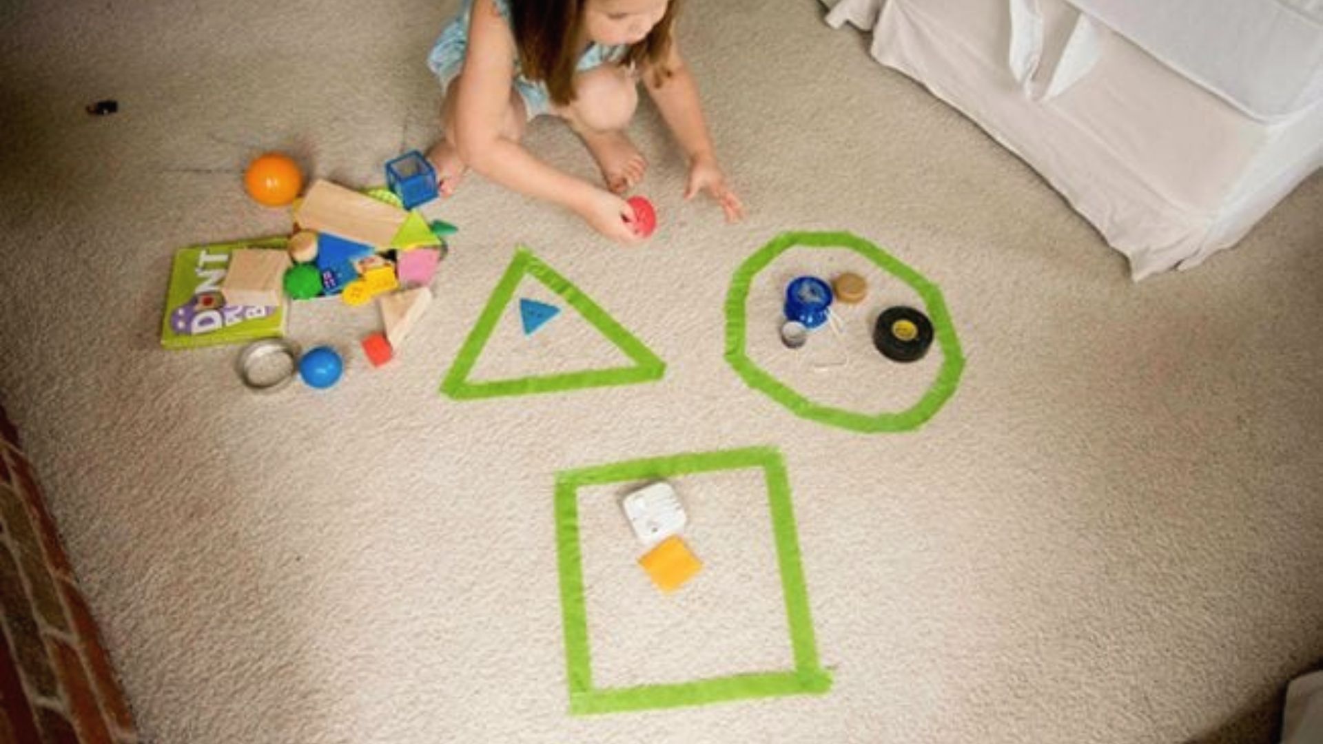 A kindergartener sorts toys into green tape outlines of a triangle, circle, and square on the floor.