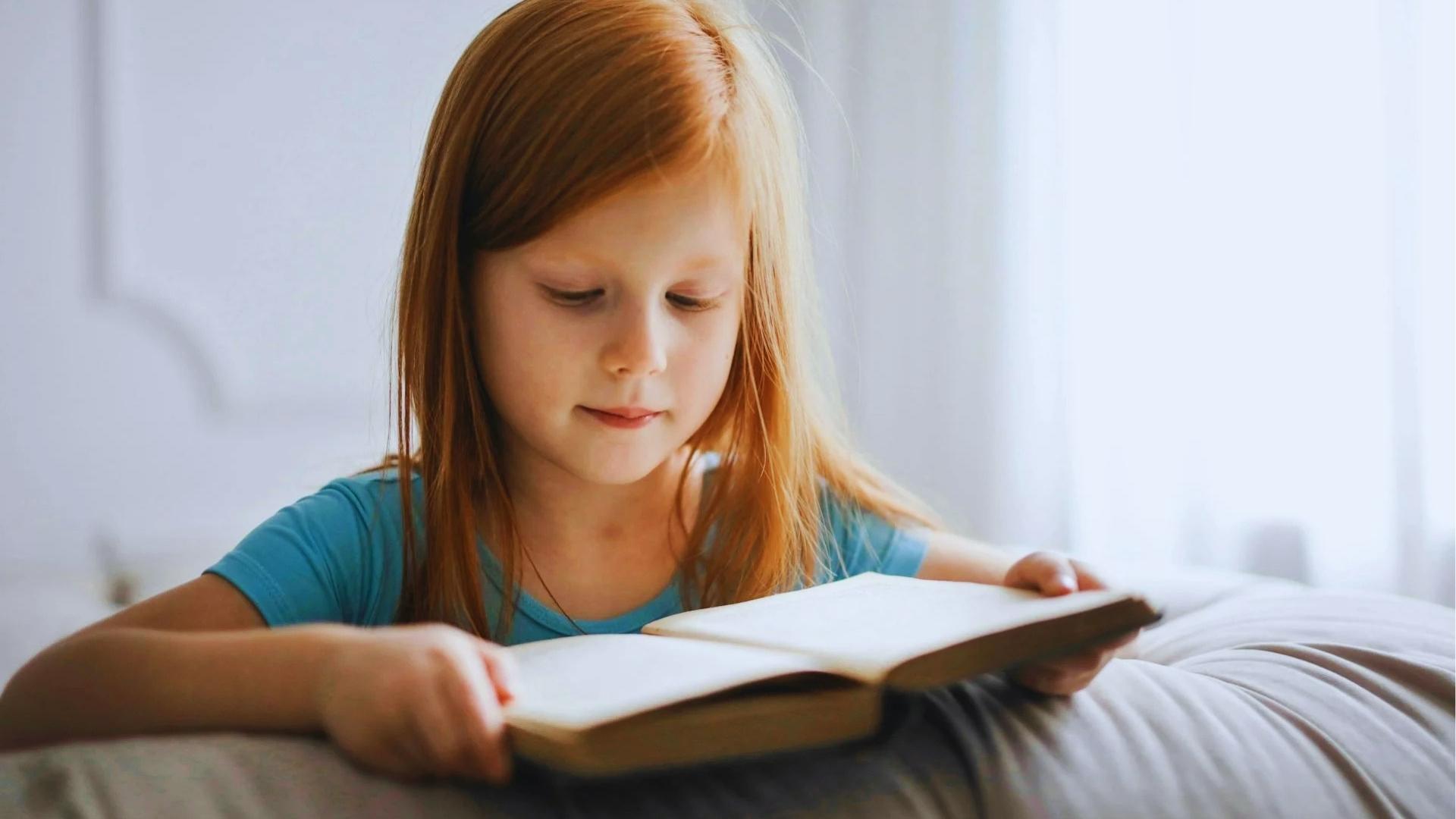 Girl in blue shirt reading chapter book independently while lying on bed in bright room.