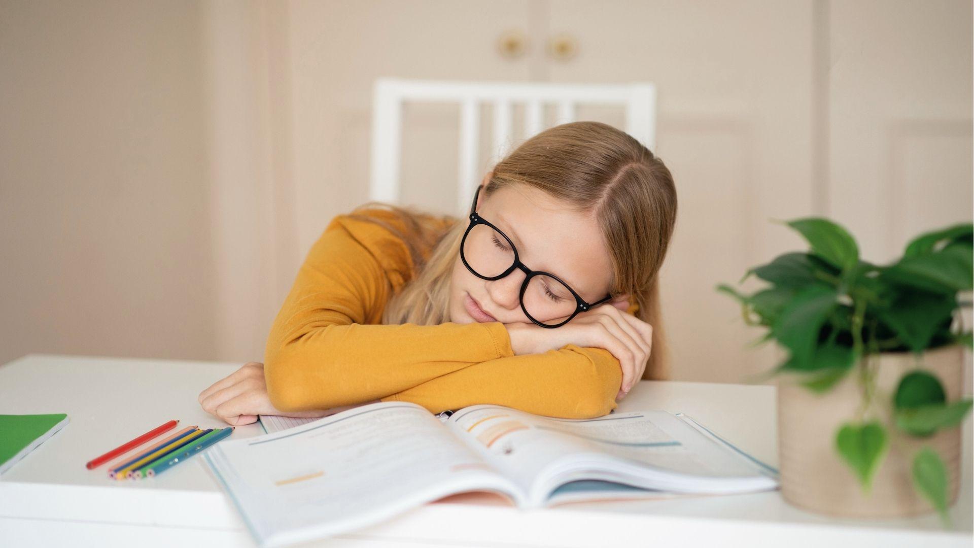 Girl in yellow shirt sleeps on open textbook at desk, showing burnout from homework procrastination.