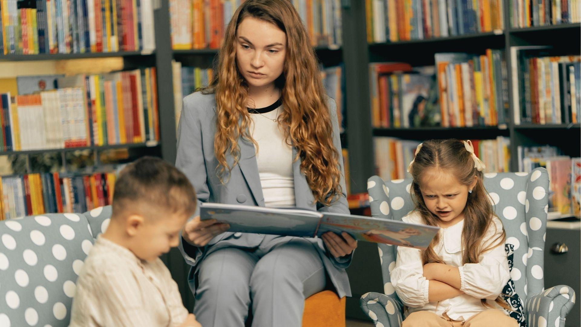 A teacher in a library reads aloud to two children, while a young girl with crossed arms looks frustrated.