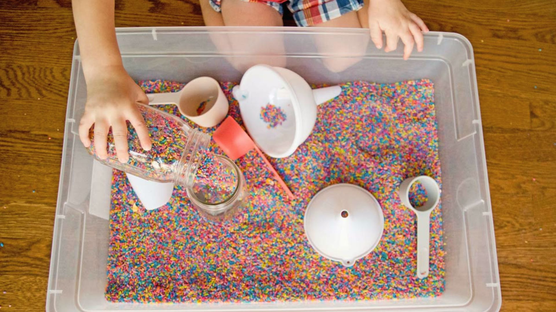 A child pours colorful rice from a jar into a sensory bin using funnels and scoops.