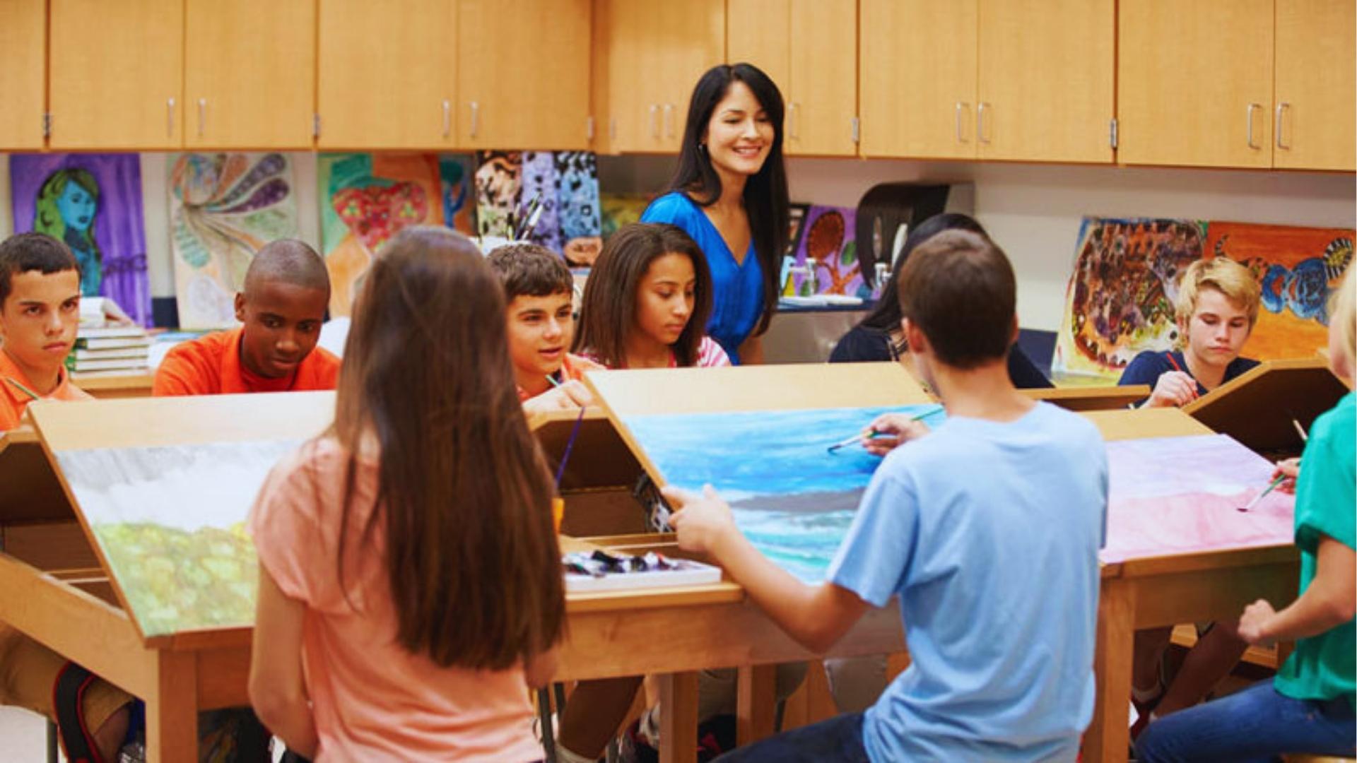 Art teacher in blue shirt guiding group of middle school students painting landscapes on easels in the classroom.