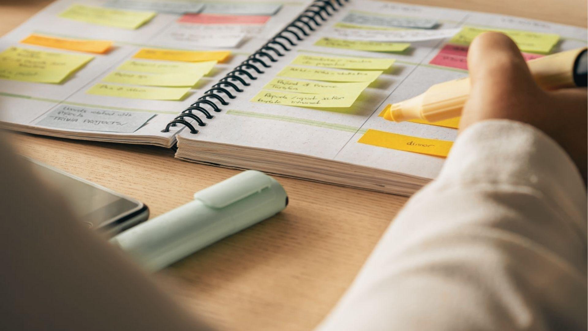 Hand holding a yellow highlighter marks a planner filled with colorful sticky notes and tasks.
