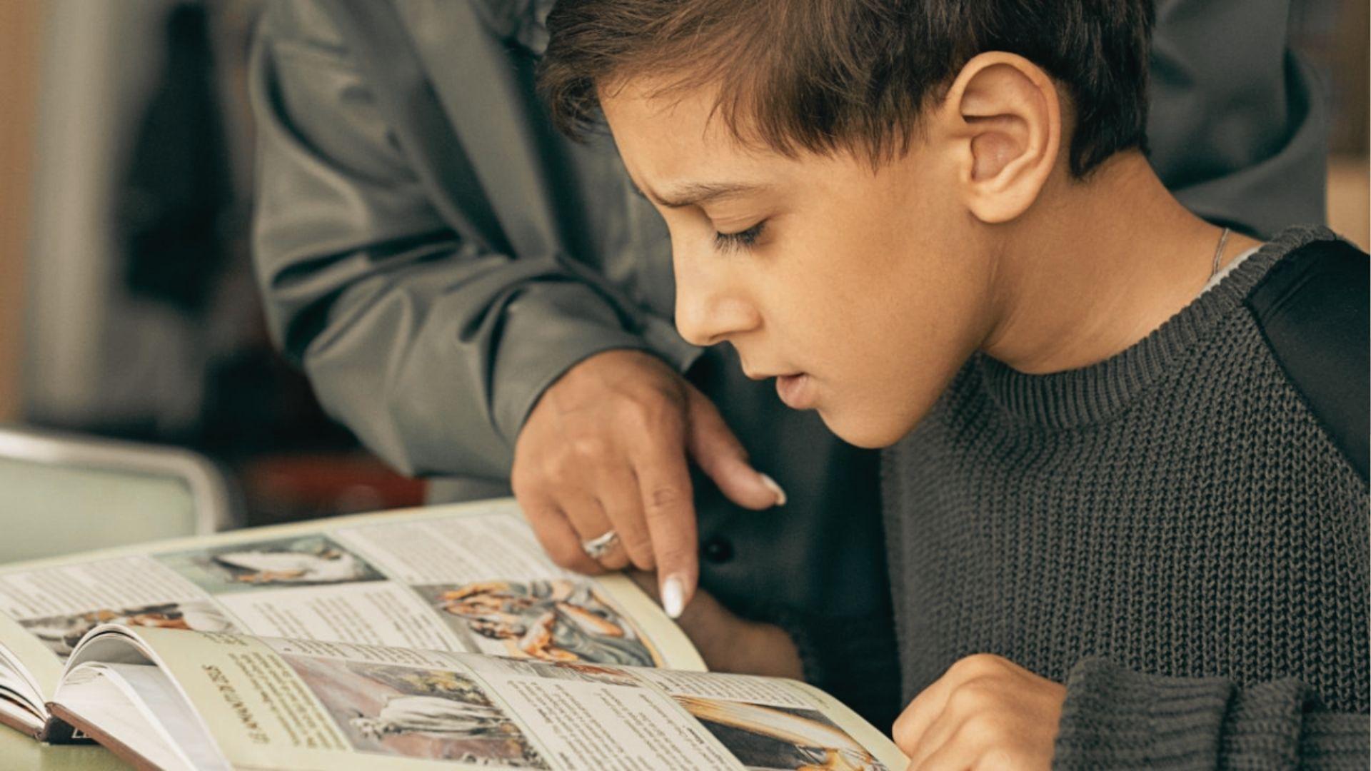 A tutor points to a textbook while a boy in a gray sweater focuses intently on the page.