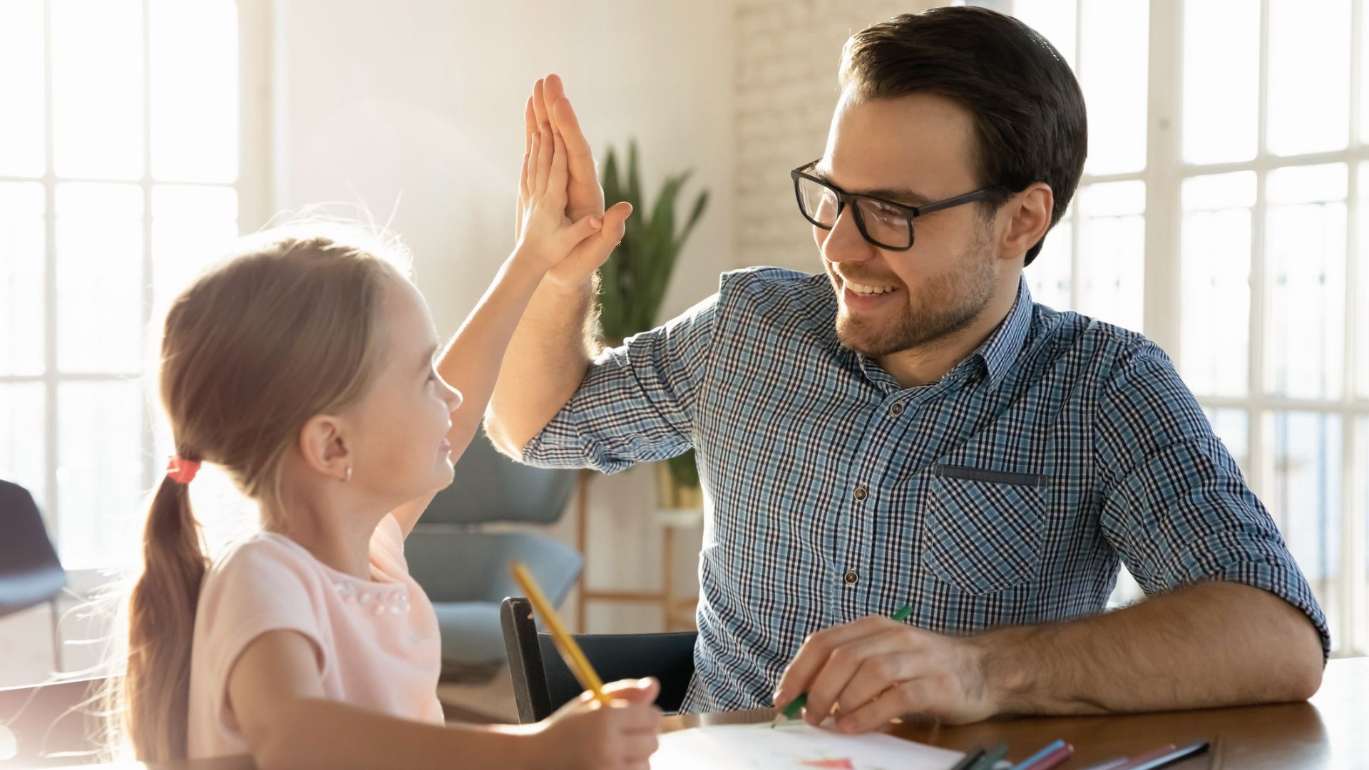 A father and daughter high-five while drawing at a bright table, both smiling with encouragement.