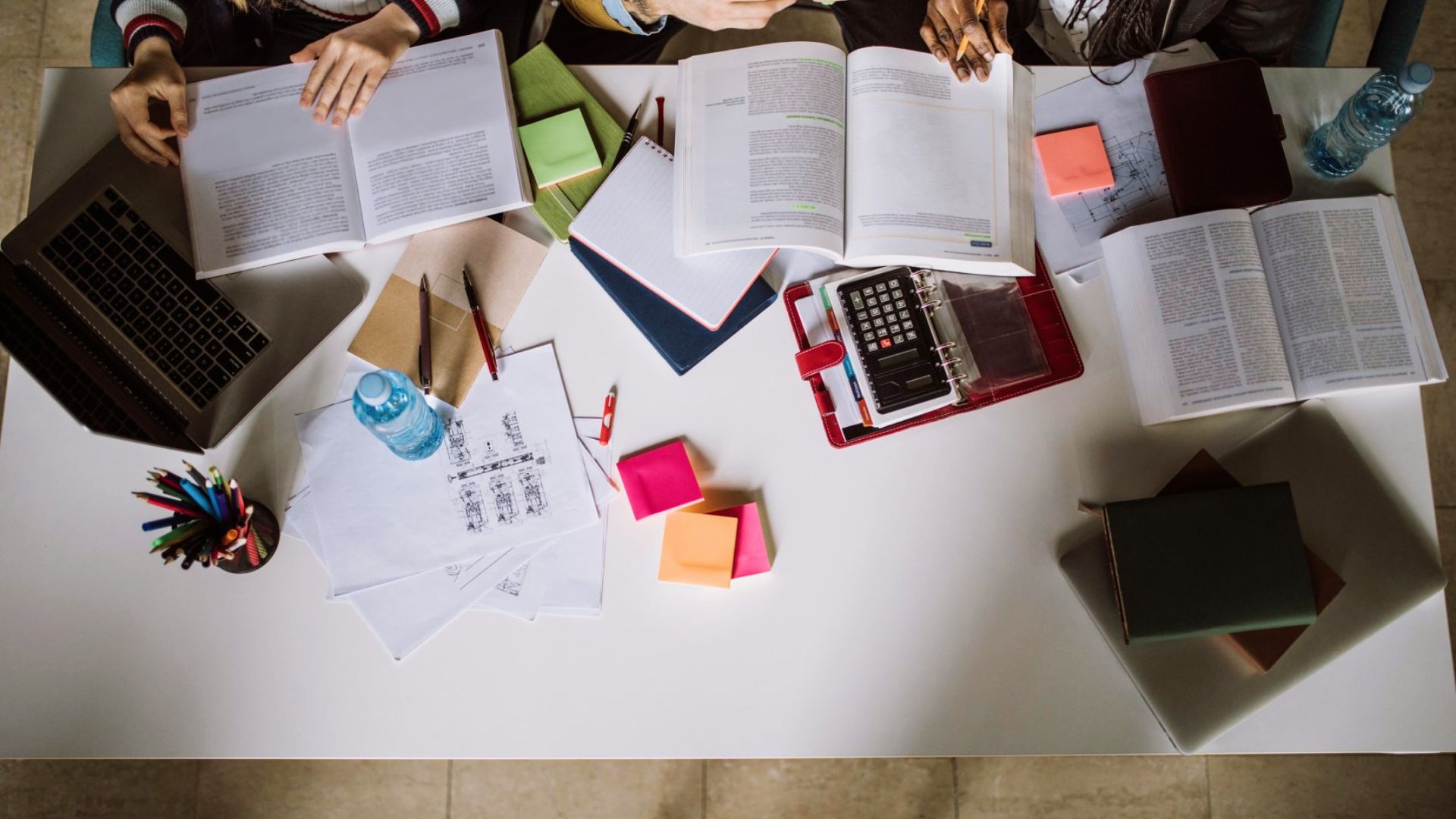 Students collaborate around a table covered with open textbooks, sticky notes, a laptop, and a calculator.