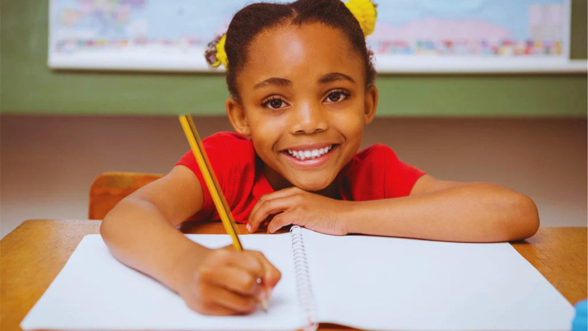 Girl in red shirt with yellow hair bows grinning at camera while writing in notebook with pencil.