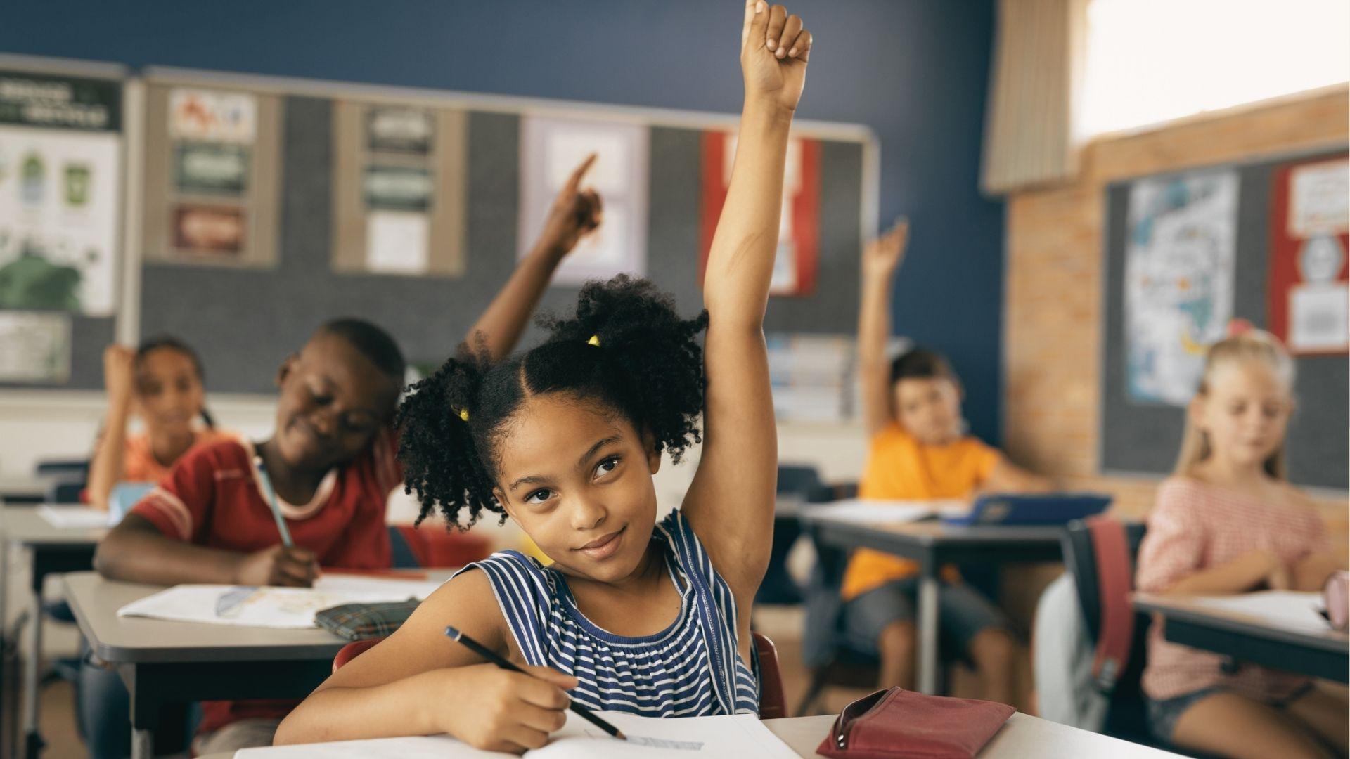 A girl with curly hair raises her hand confidently as other students do the same in a bright classroom.