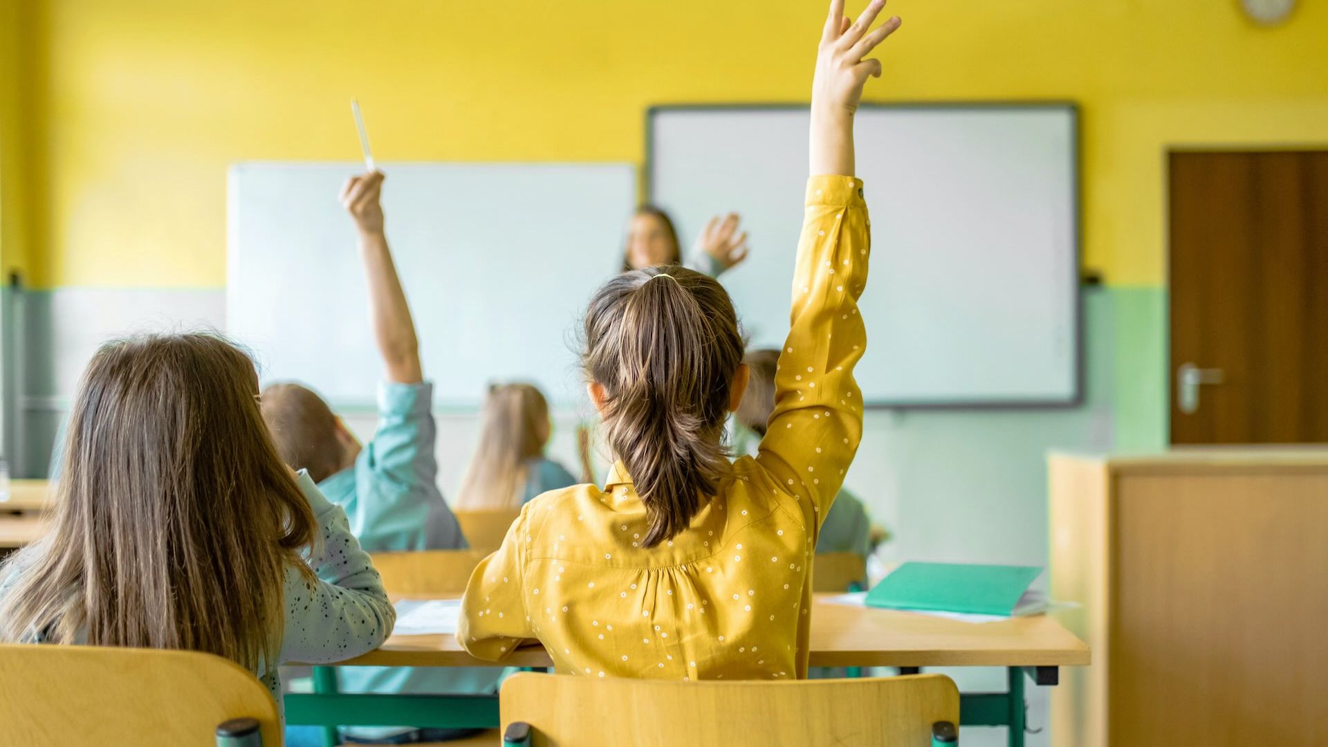 Students raise their hands to participate in a classroom discussion with a teacher at the board.