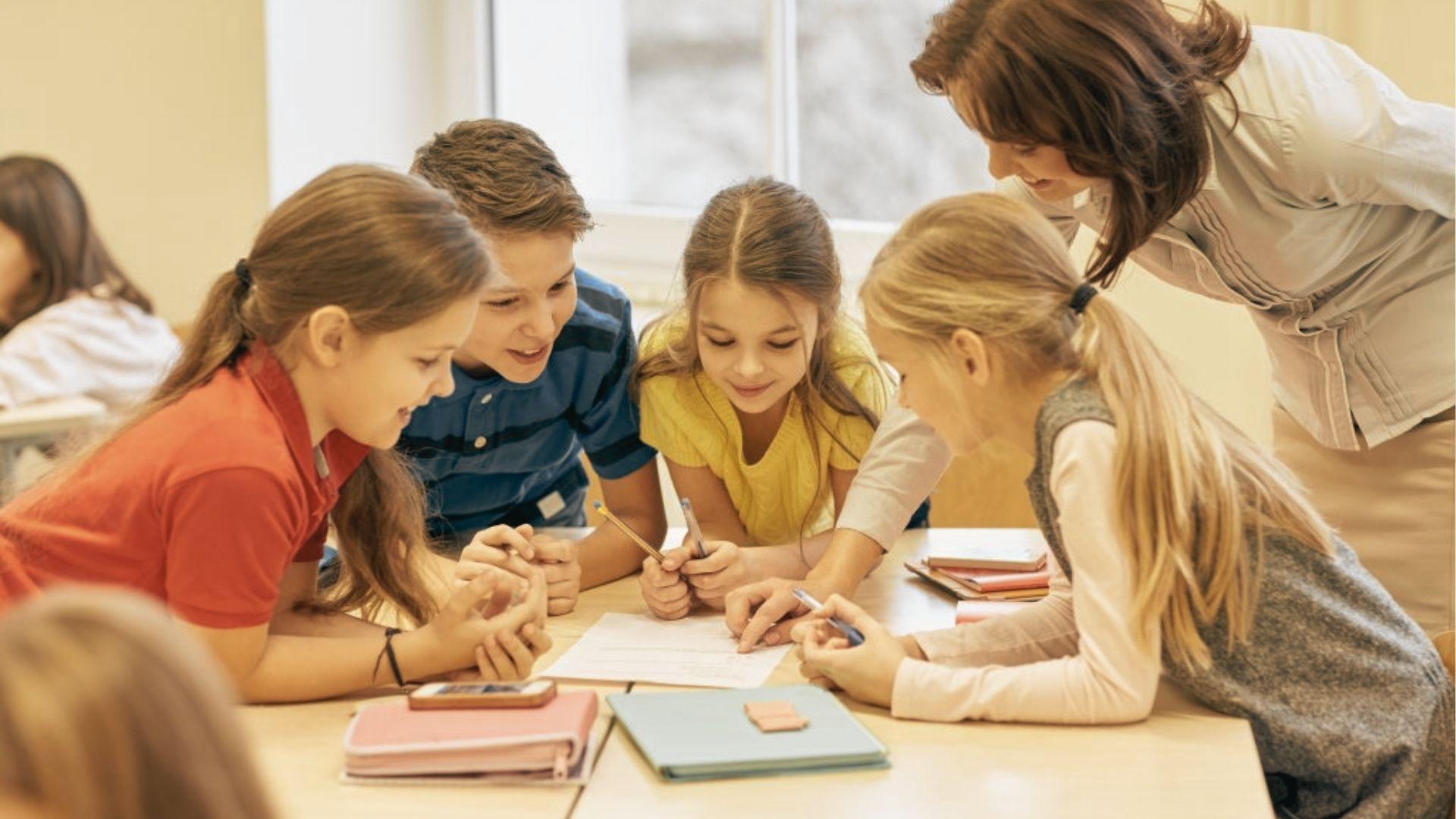 A teacher leans over a table while four students huddle around a shared paper, pointing and discussing together.