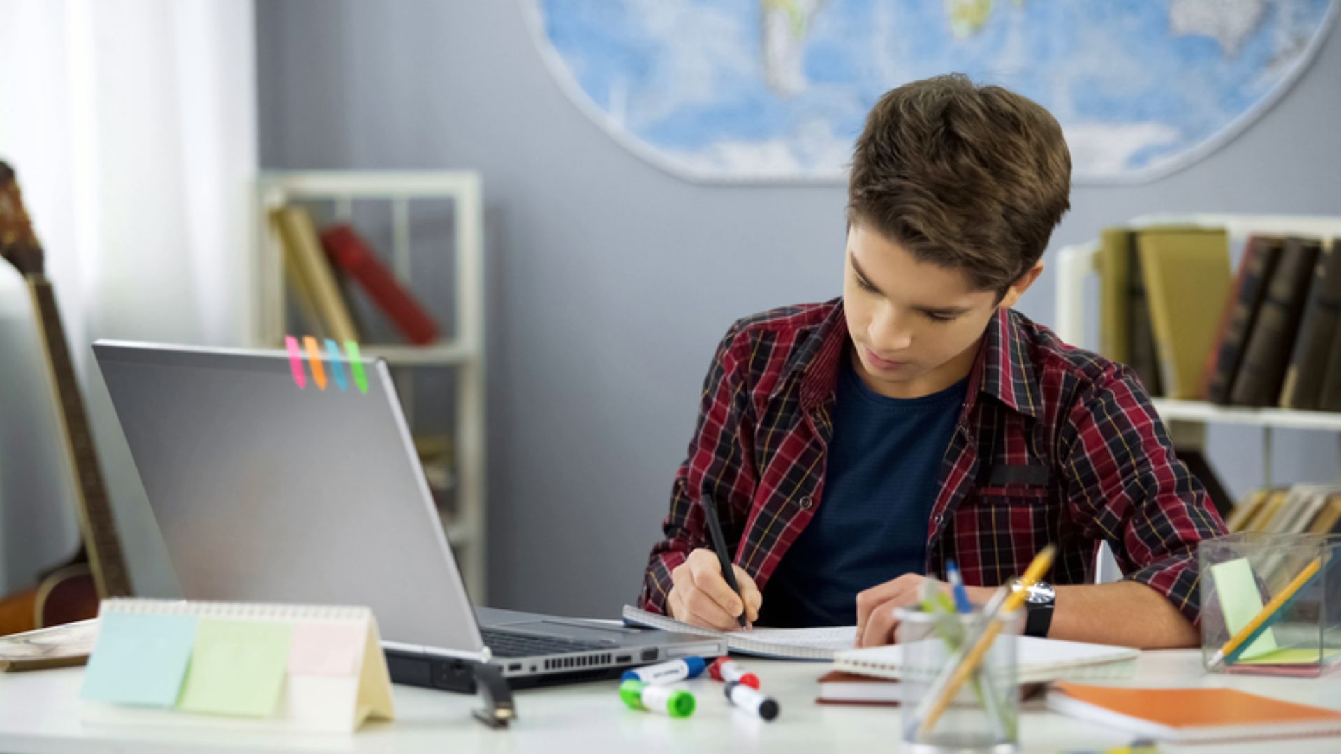 A teen boy takes handwritten notes at a neat desk with a laptop, sticky notes, and markers.