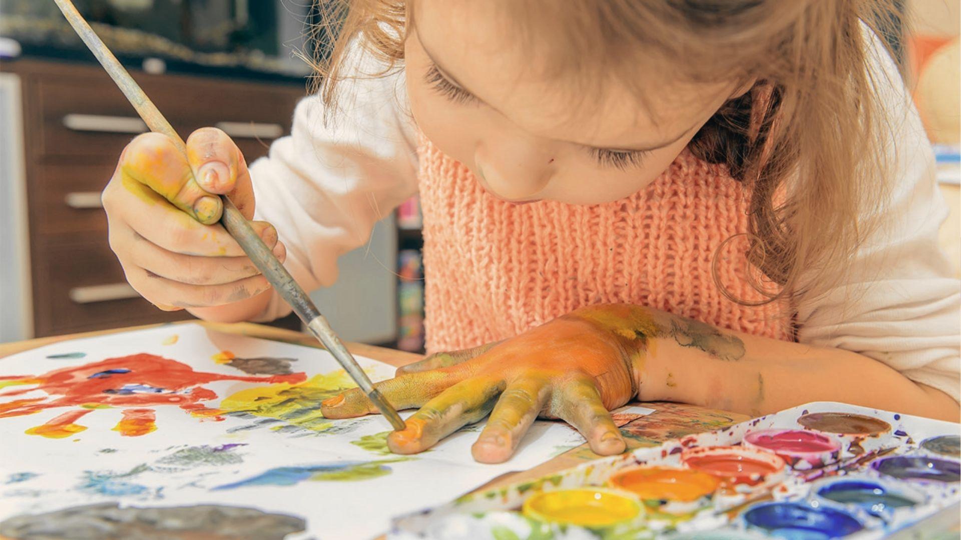 A preschooler in a peach sweater paints their own hand with a brush while working on colorful handprint art at a table.