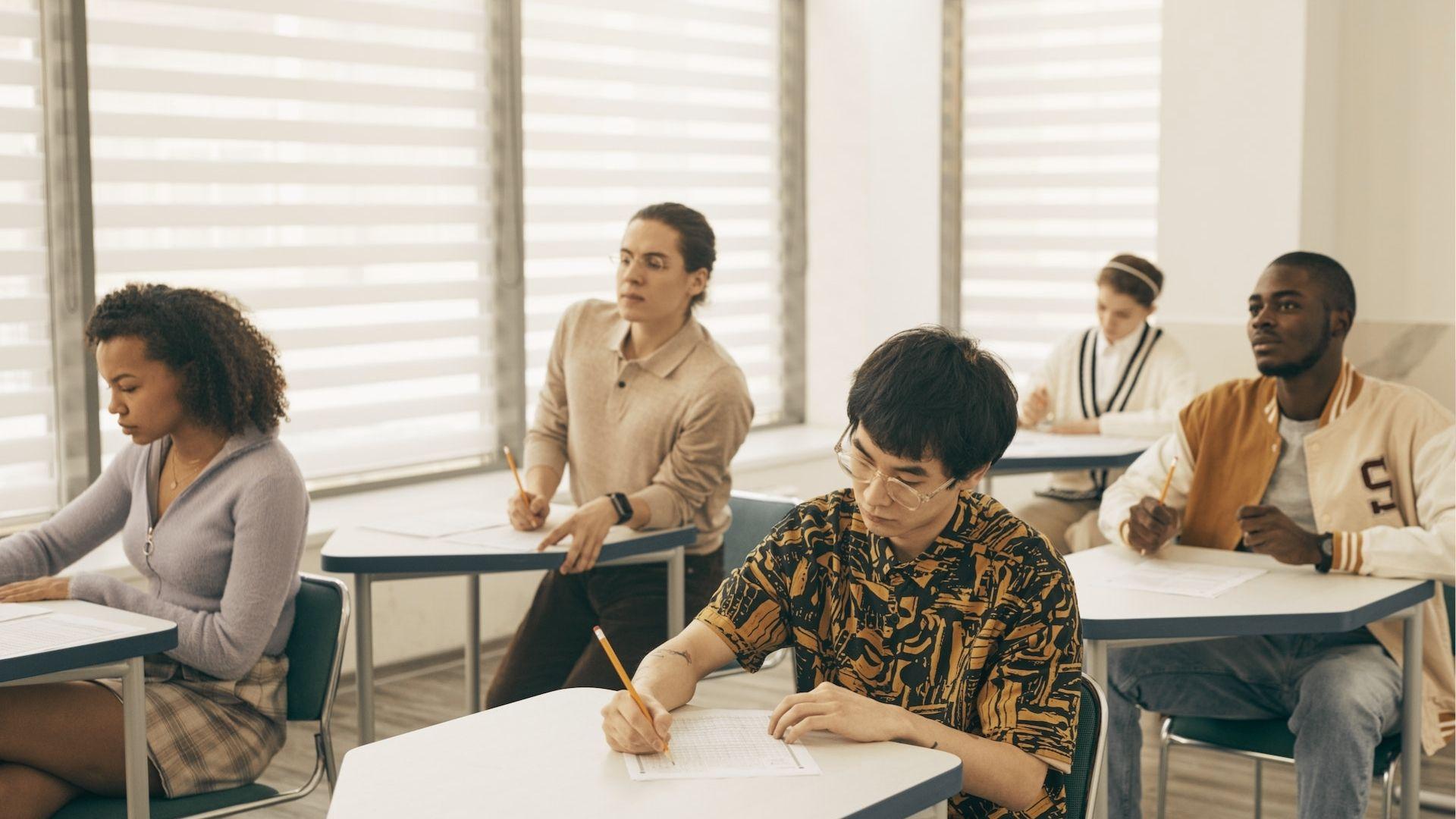 Diverse group of high school students takes a test at individual desks in a sunlit classroom.