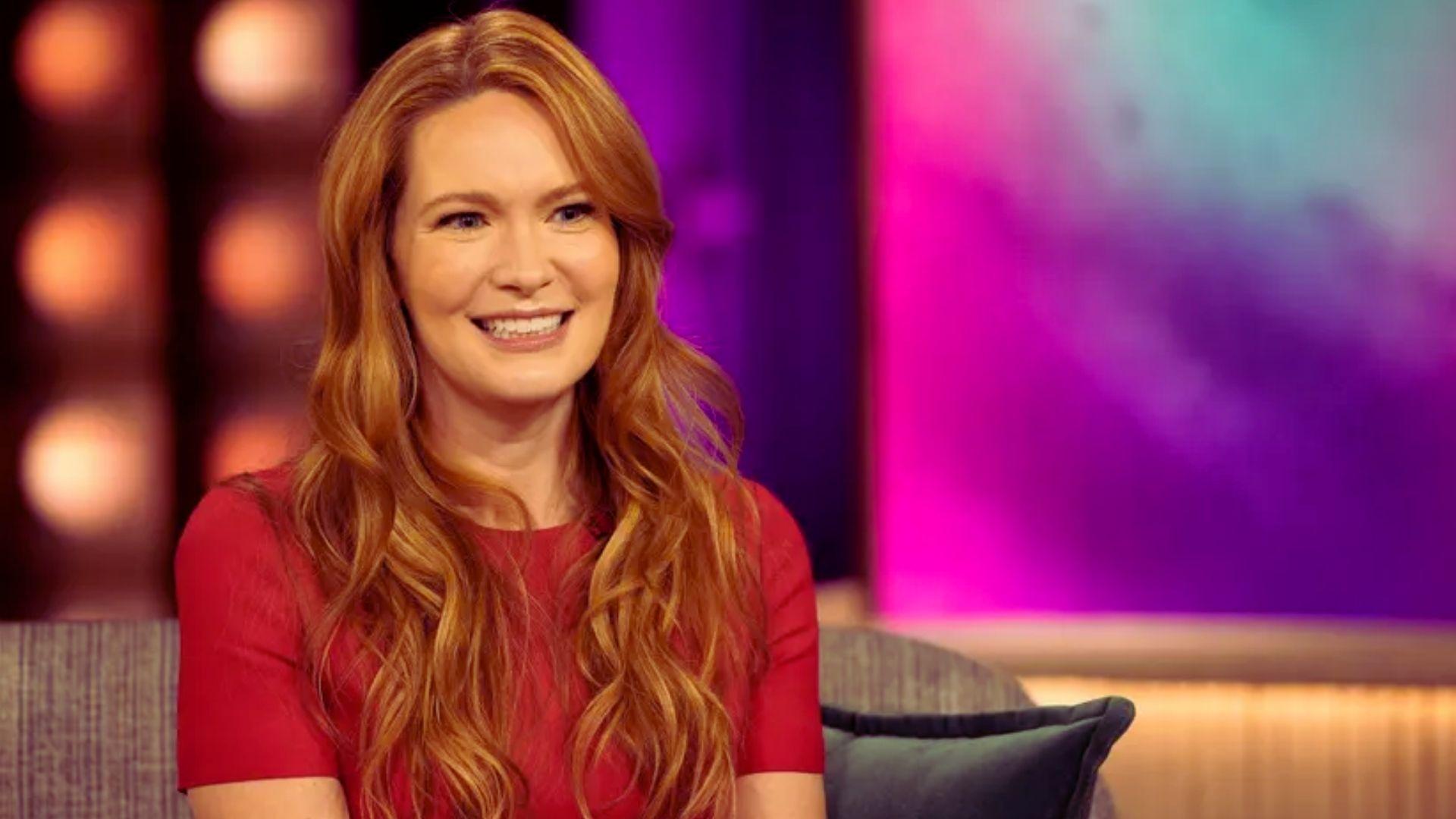 A woman with long red hair is sitting comfortably on a couch, looking relaxed and engaged in her surroundings.