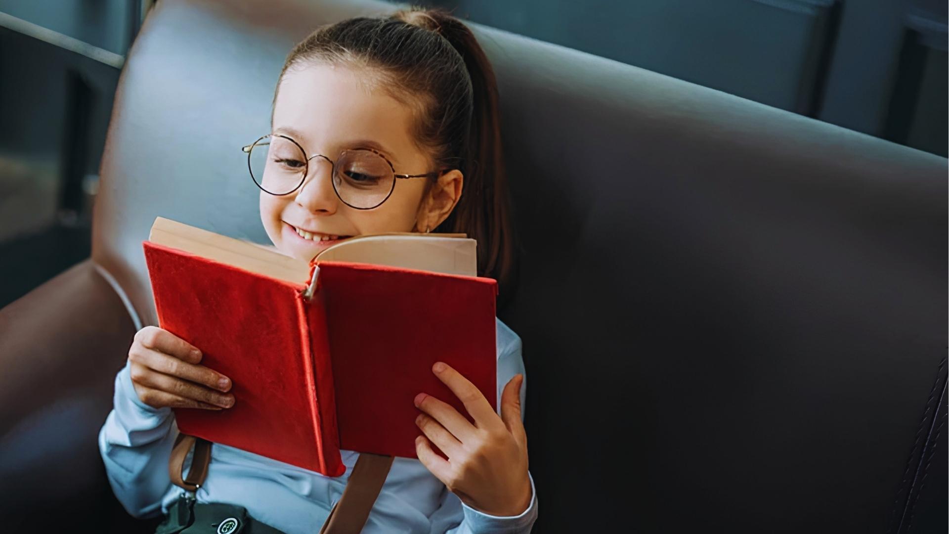 Girl with round glasses and ponytail smiling while reading red hardcover book on brown leather sofa.