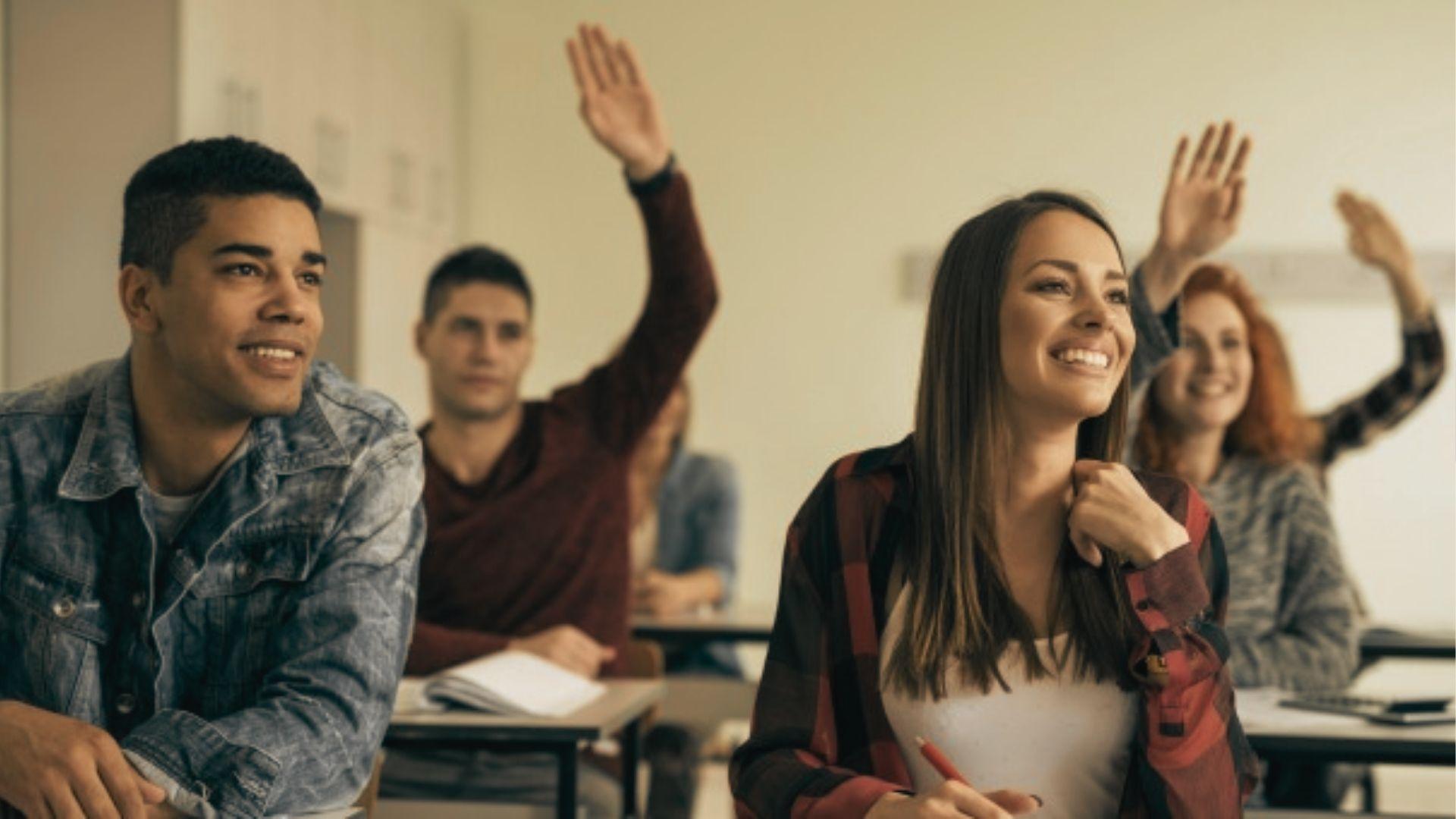 Smiling high school students raise their hands to participate in a bright classroom.