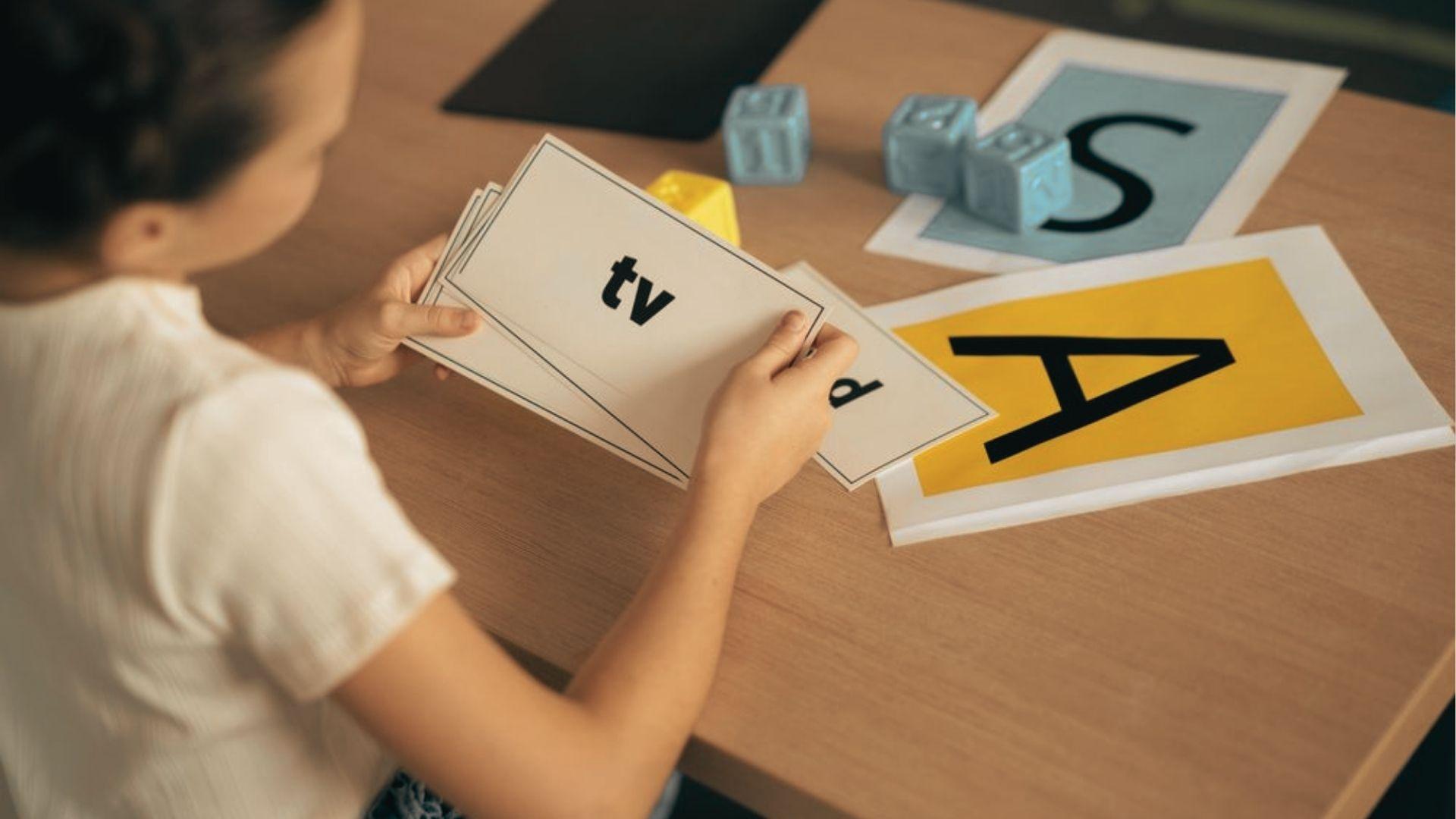 A child holds flashcards with the word "tv" while letter cards and blocks for A and S sit on the table.