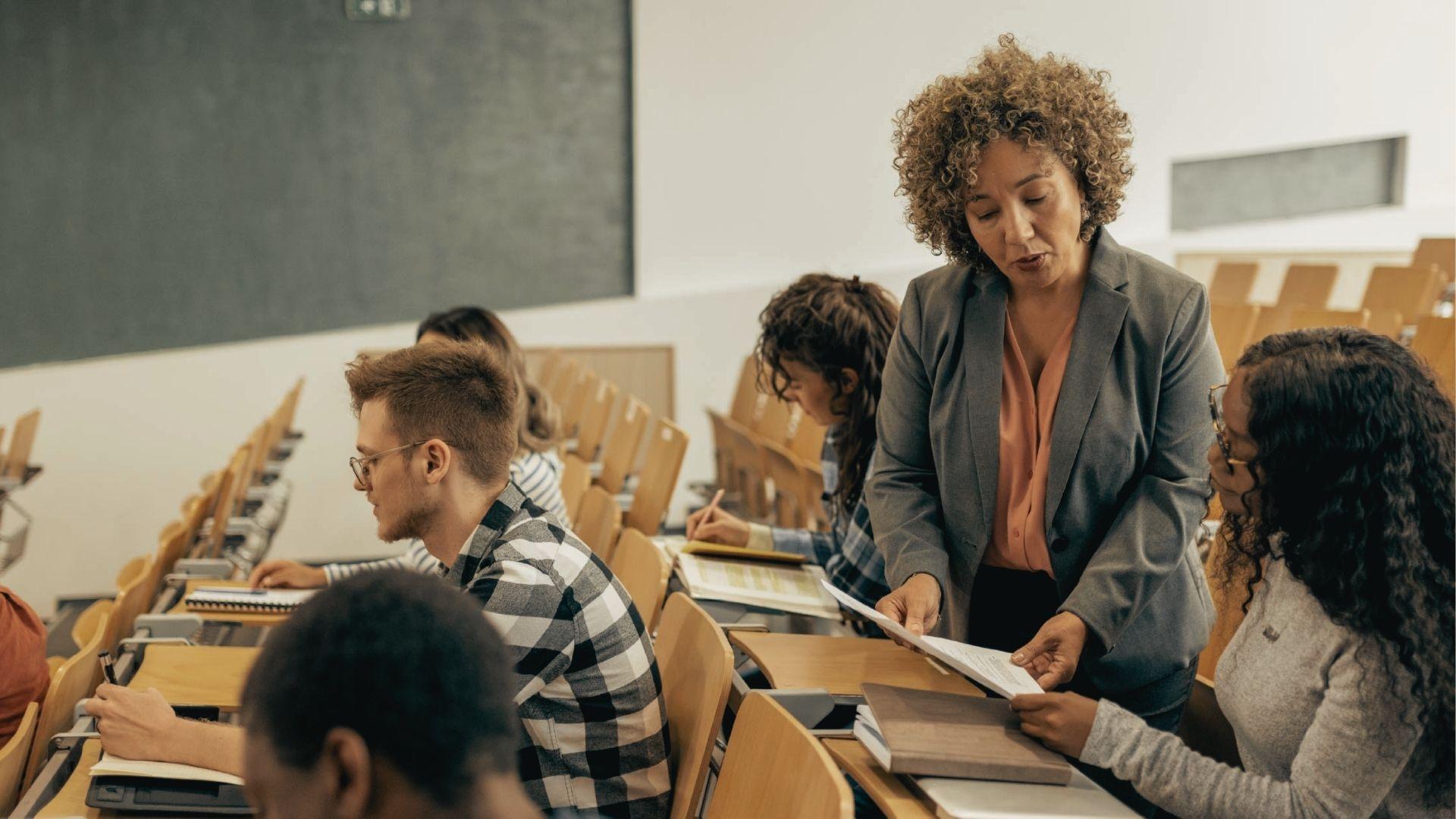Teacher leaning over a desk to help a student with her paper in a lecture hall.