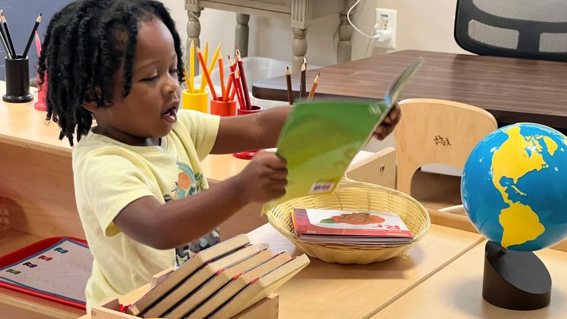 A kindergartener finds picture books at a classroom table beside a globe and colored pencils.