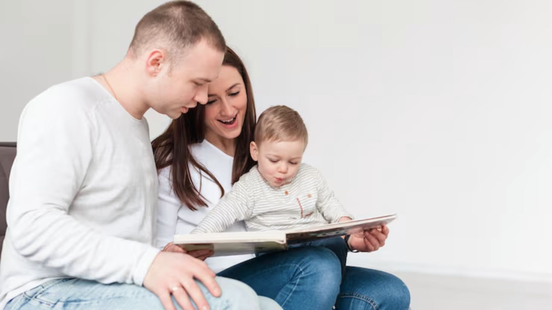 Parents and their toddler sit close, reading a book together with happy, engaged expressions.