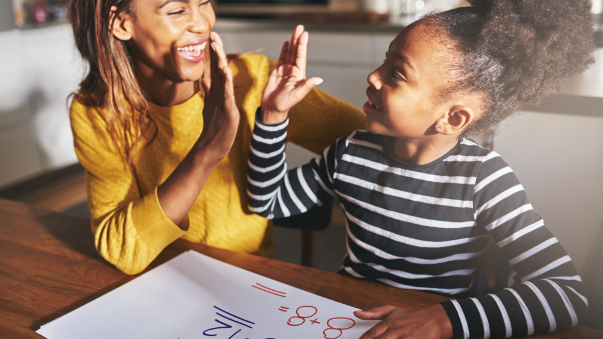 A smiling mother and daughter share a high-five over math homework at a sunlit kitchen table.