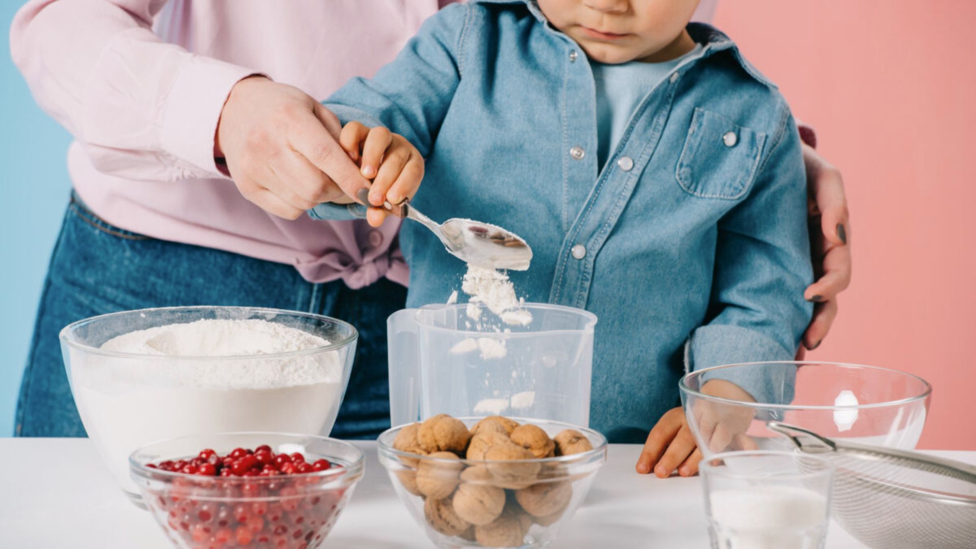 An adult guides a child’s hand as they scoop flour into a container with other baking ingredients.