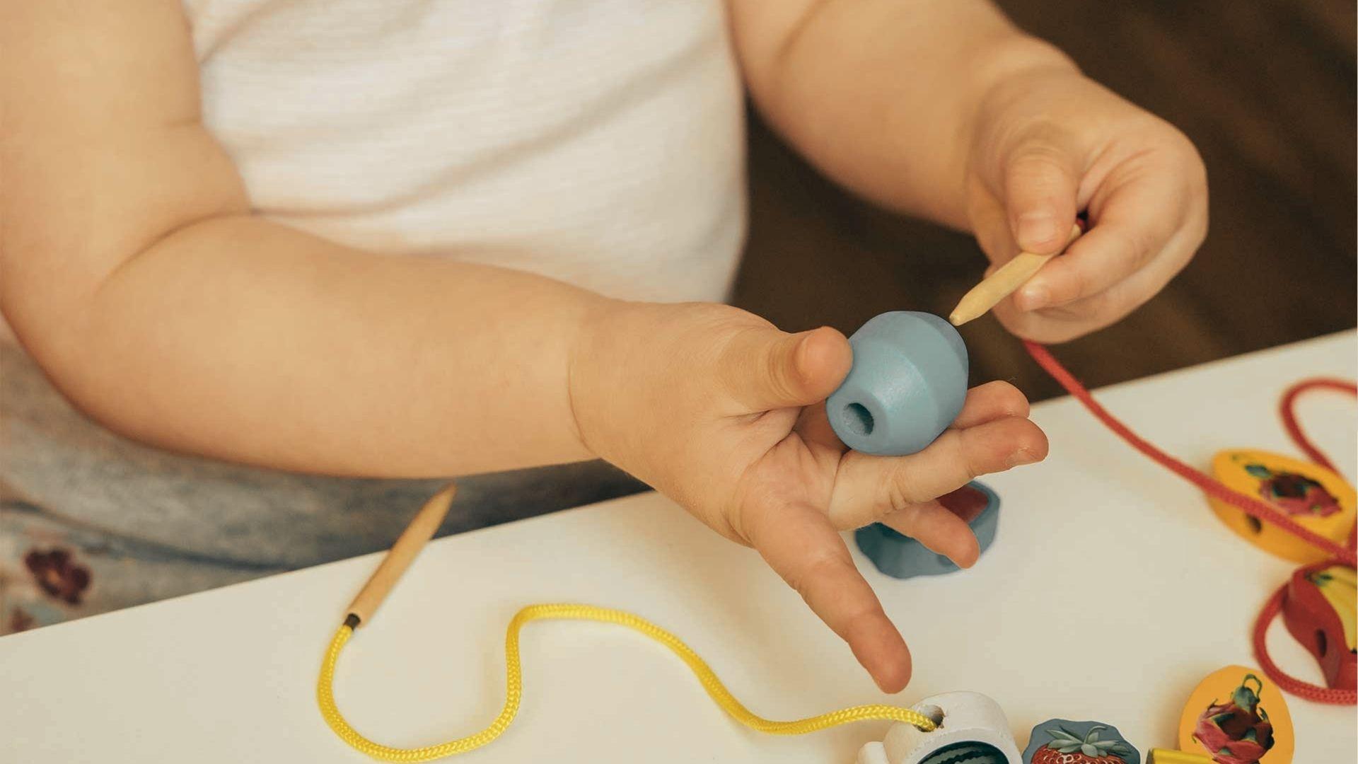 A young child threads a blue wooden bead onto a red string using a wooden needle and steady hands.