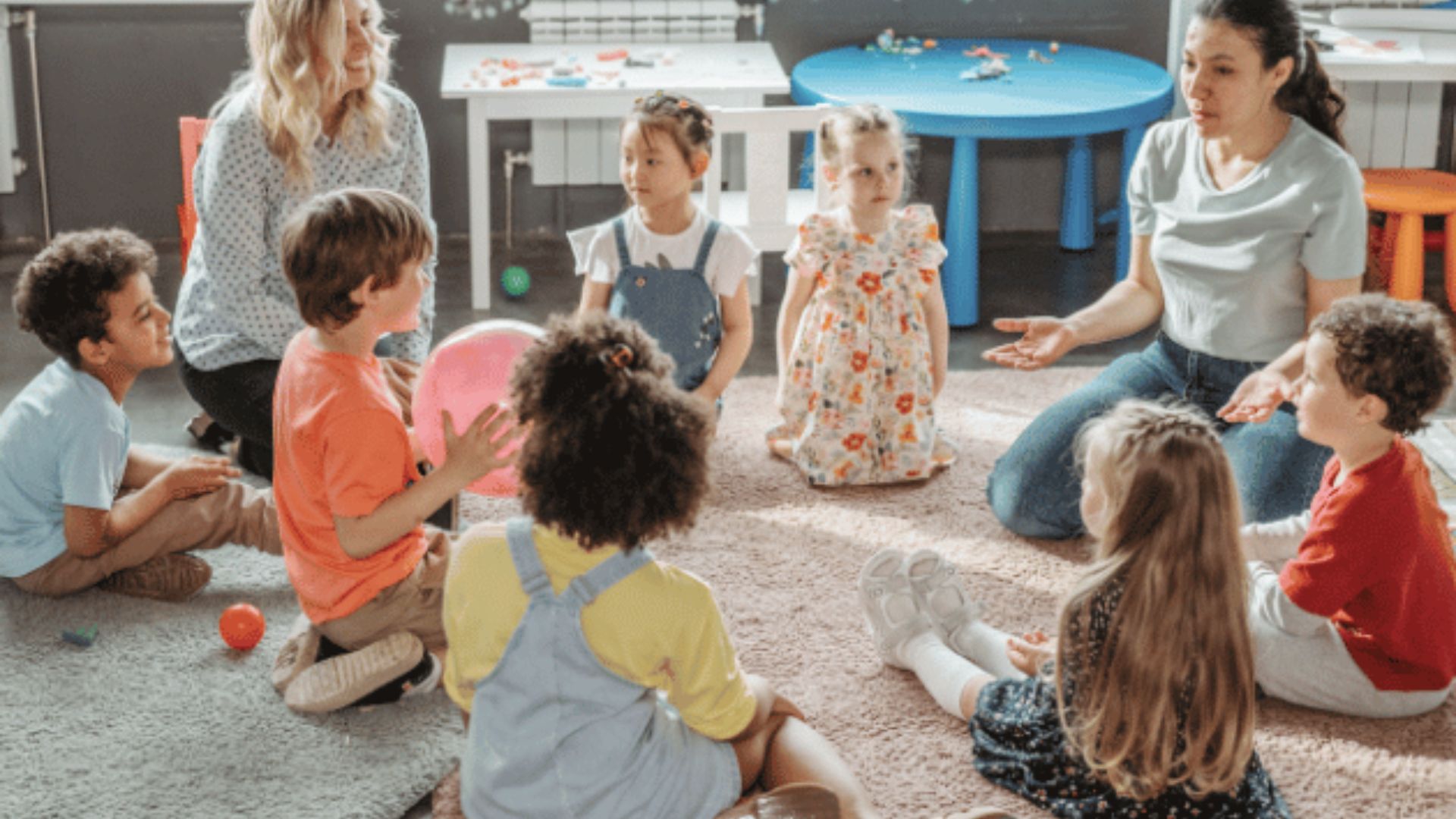 Two teachers sit on a classroom rug with a diverse group of young children gathered in a circle doing rhyming word basket toss.