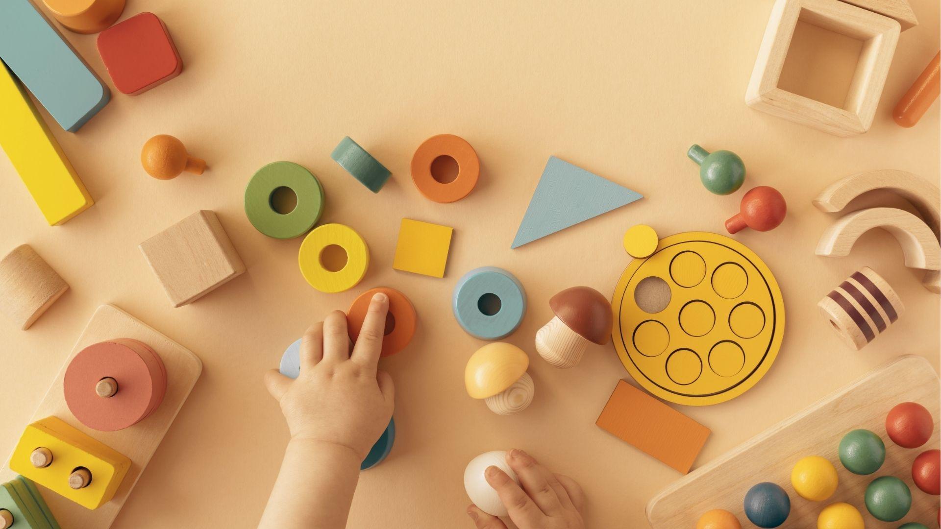 Toddler hands reach for colorful wooden shapes and stacking rings spread across a peach table.