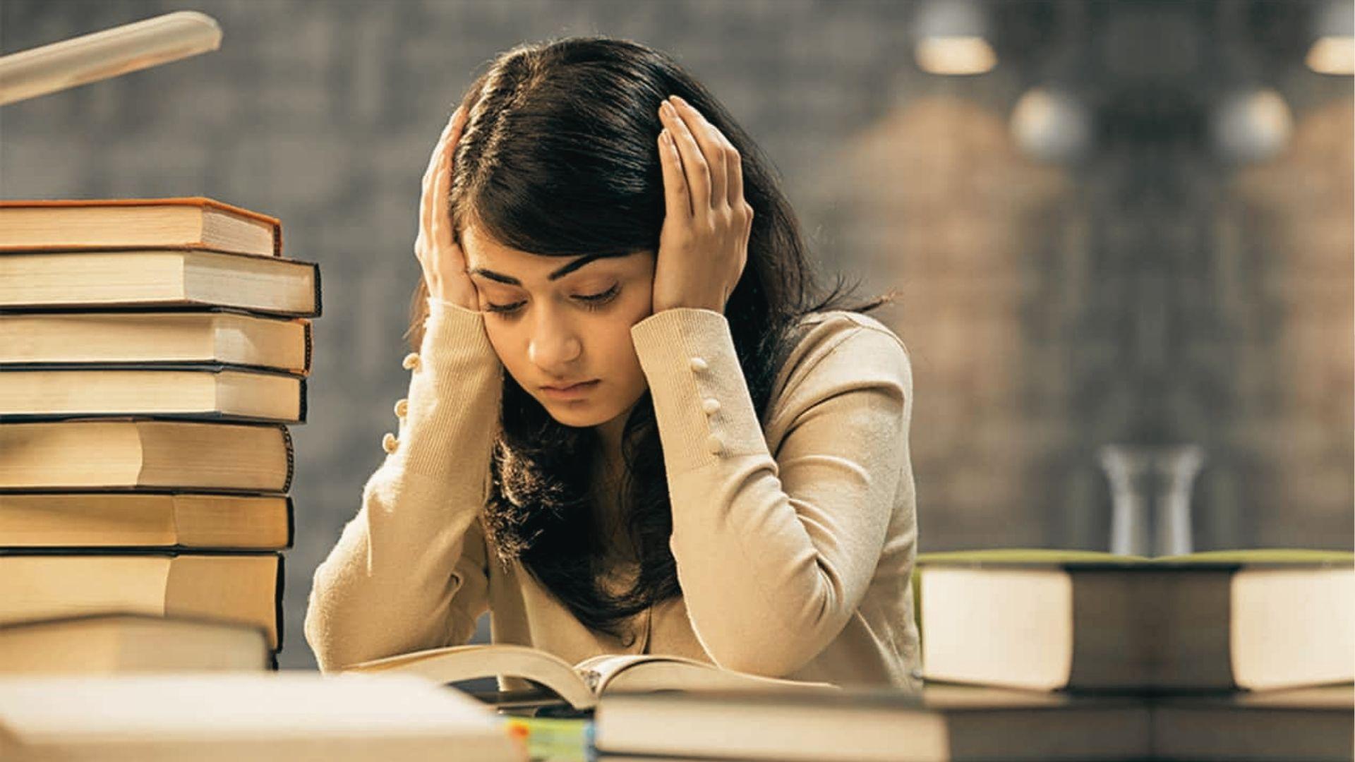 Overwhelmed female student holds her head in her hands, surrounded by tall stacks of textbooks under a desk lamp.