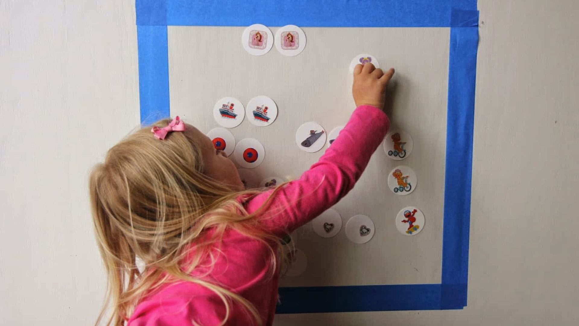 A girl in a pink shirt places round picture cards inside a taped square on the wall.