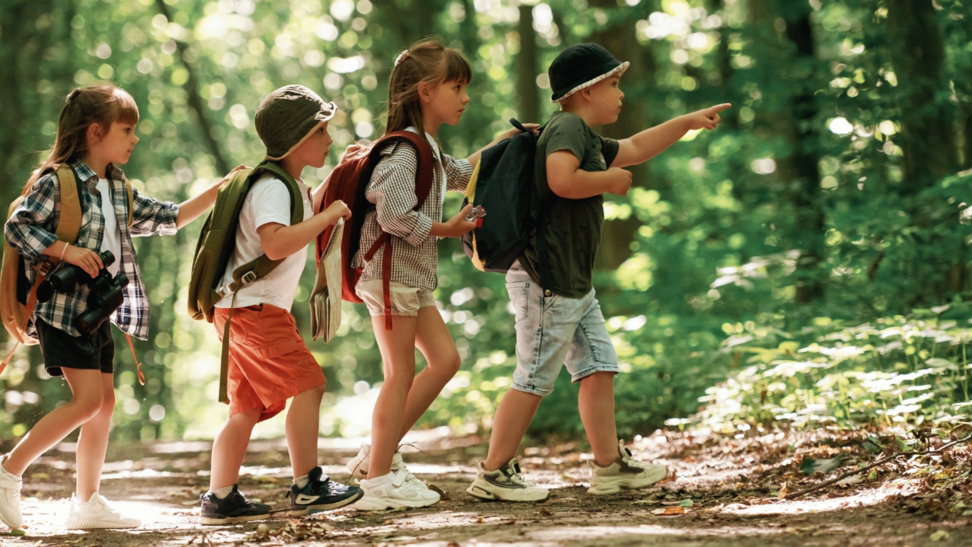 Kindergarteners with backpacks and binoculars walk in a forest line as one child points ahead.