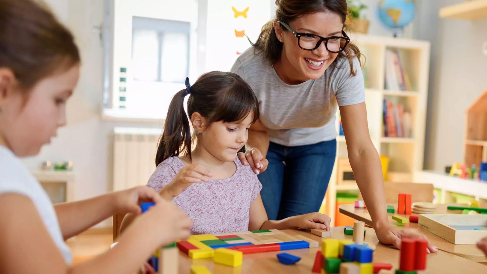 A teacher smiles as she guides kindergarteners building patterns with colorful wooden blocks.