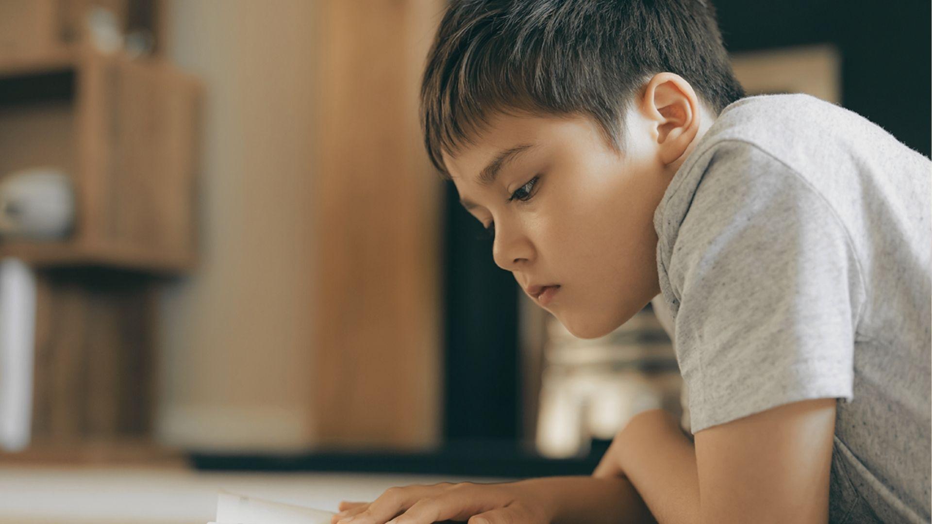 A young boy leans over a book, eyes focused and lips slightly parted in concentration.