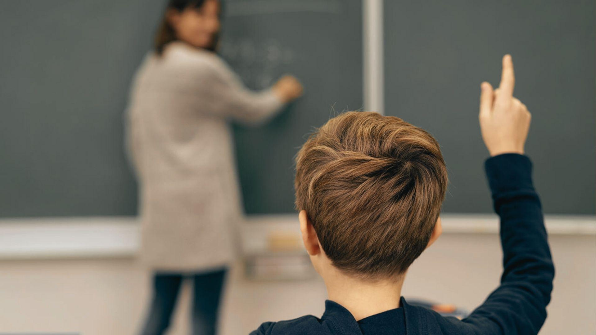 A student raises his hand in class while a teacher writes on the chalkboard in the background.