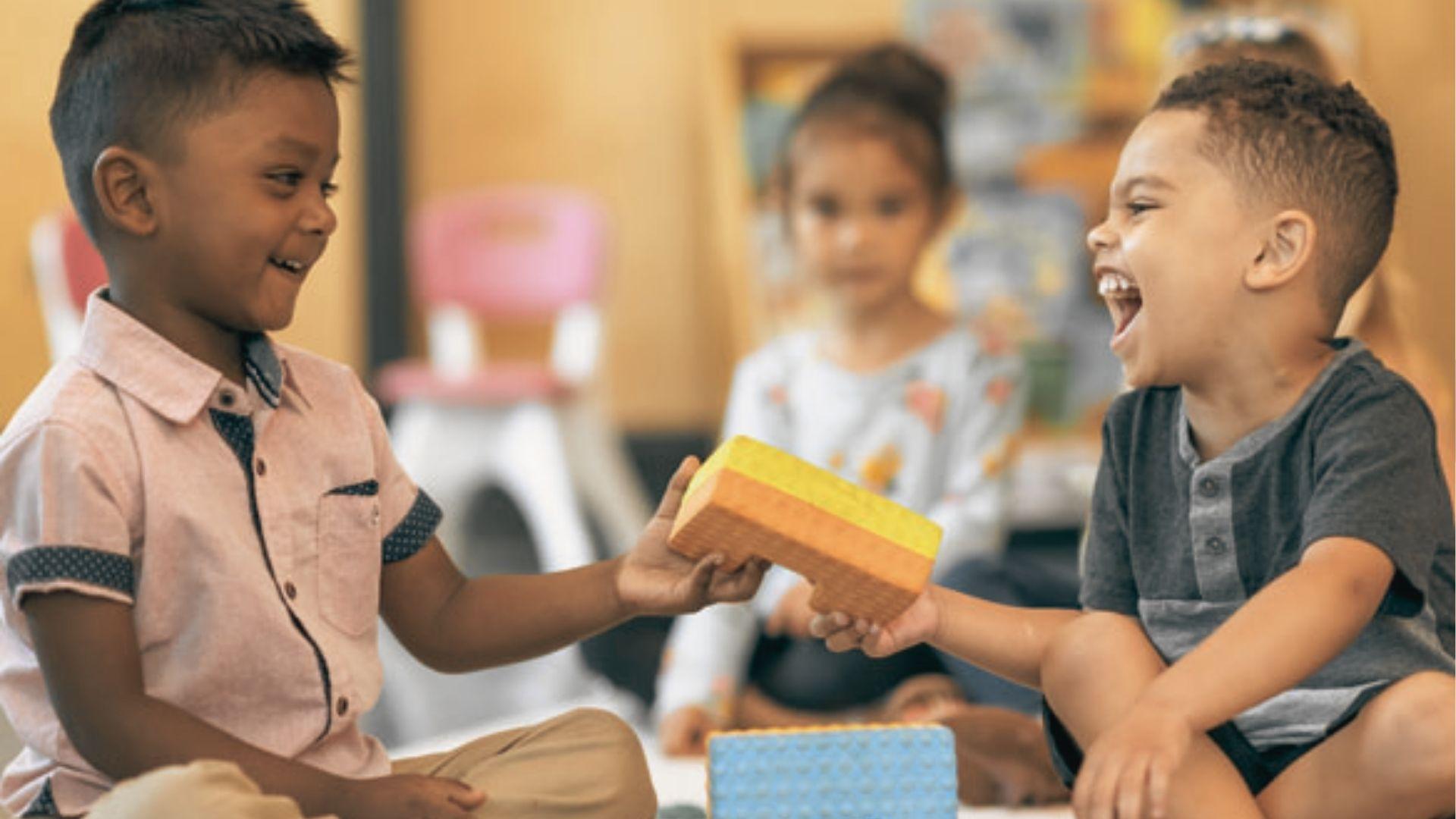 Two preschool boys sit facing each other, laughing and sharing a yellow foam block during playtime.