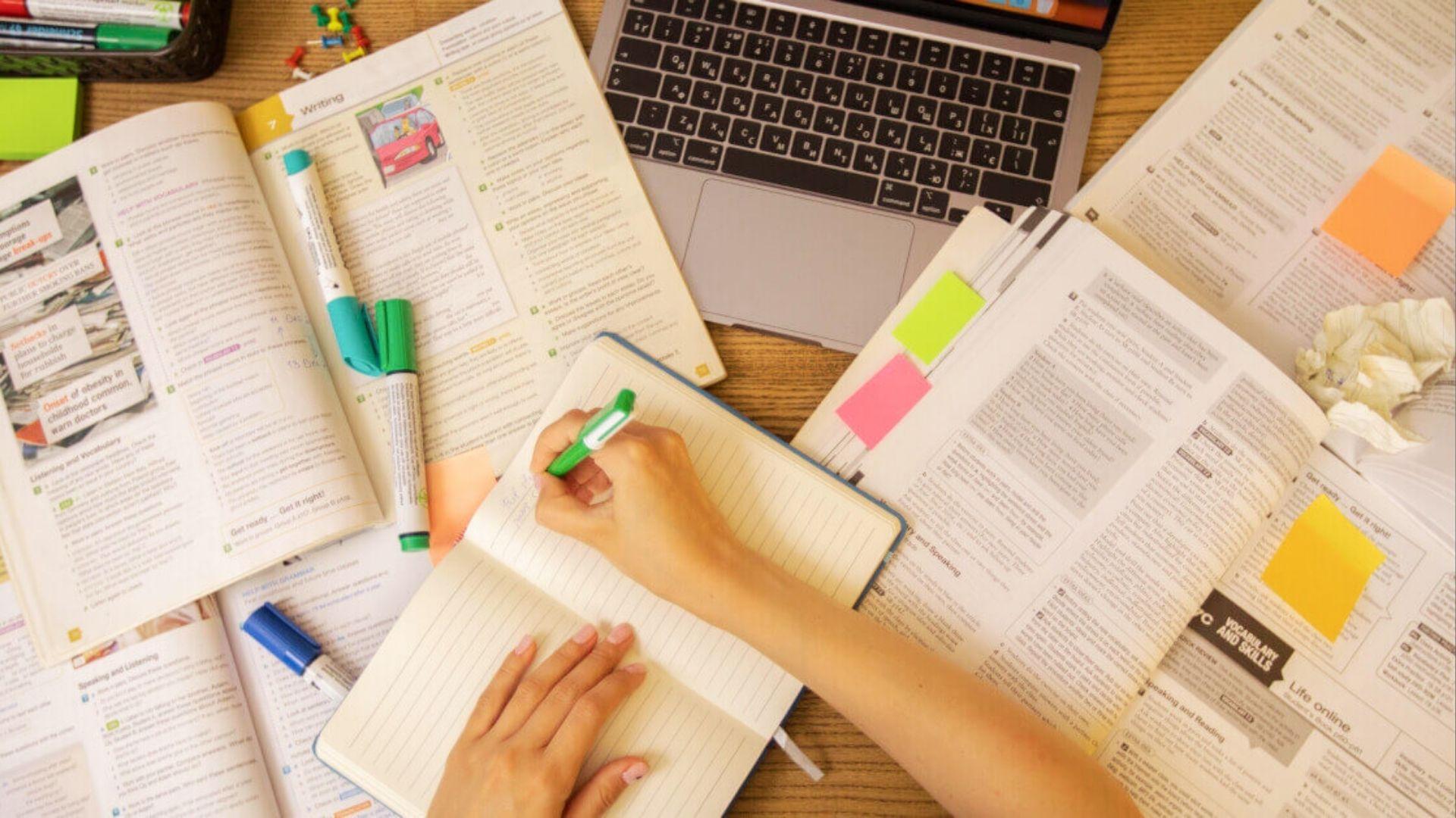 Student handwriting spelling words in notebook with open textbooks, laptop, and markers on desk