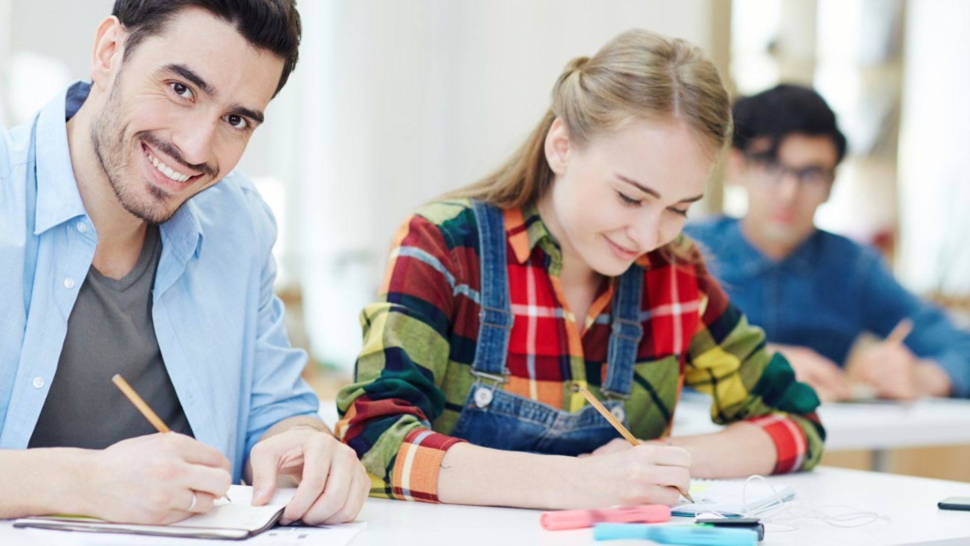 Two students smile while taking notes together in a bright classroom.
