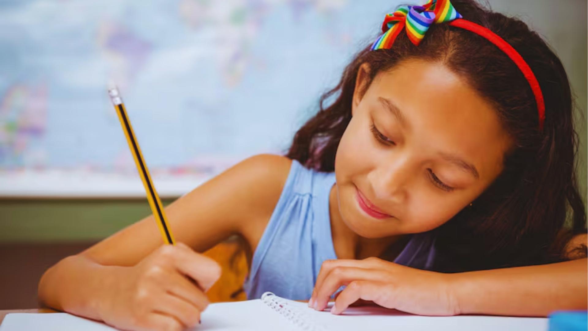 Girl with rainbow bow headband smiling while writing in spiral notebook with yellow pencil at desk.