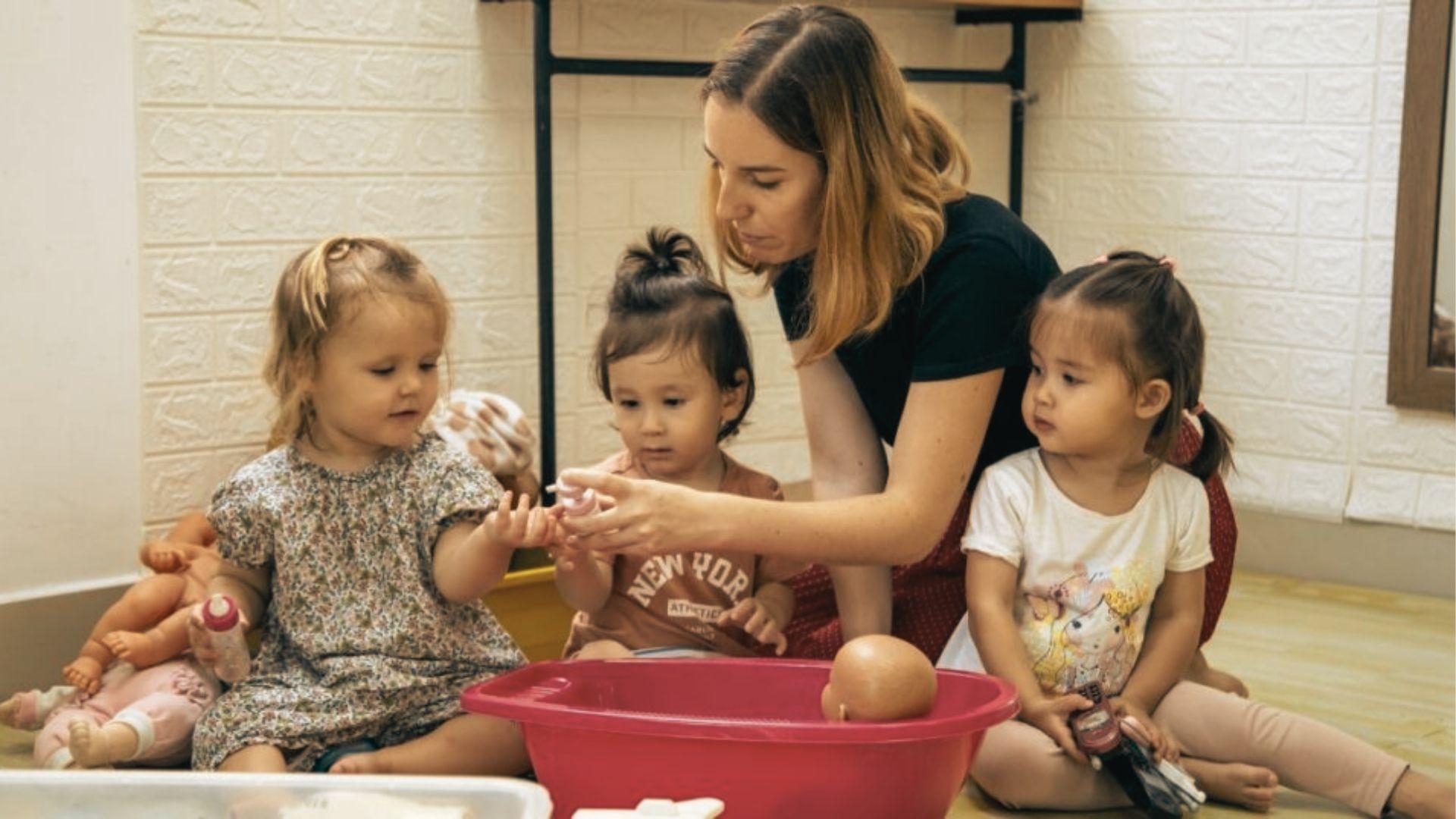 A caregiver kneels with three toddlers during pretend play, helping them care for baby dolls in a red tub.