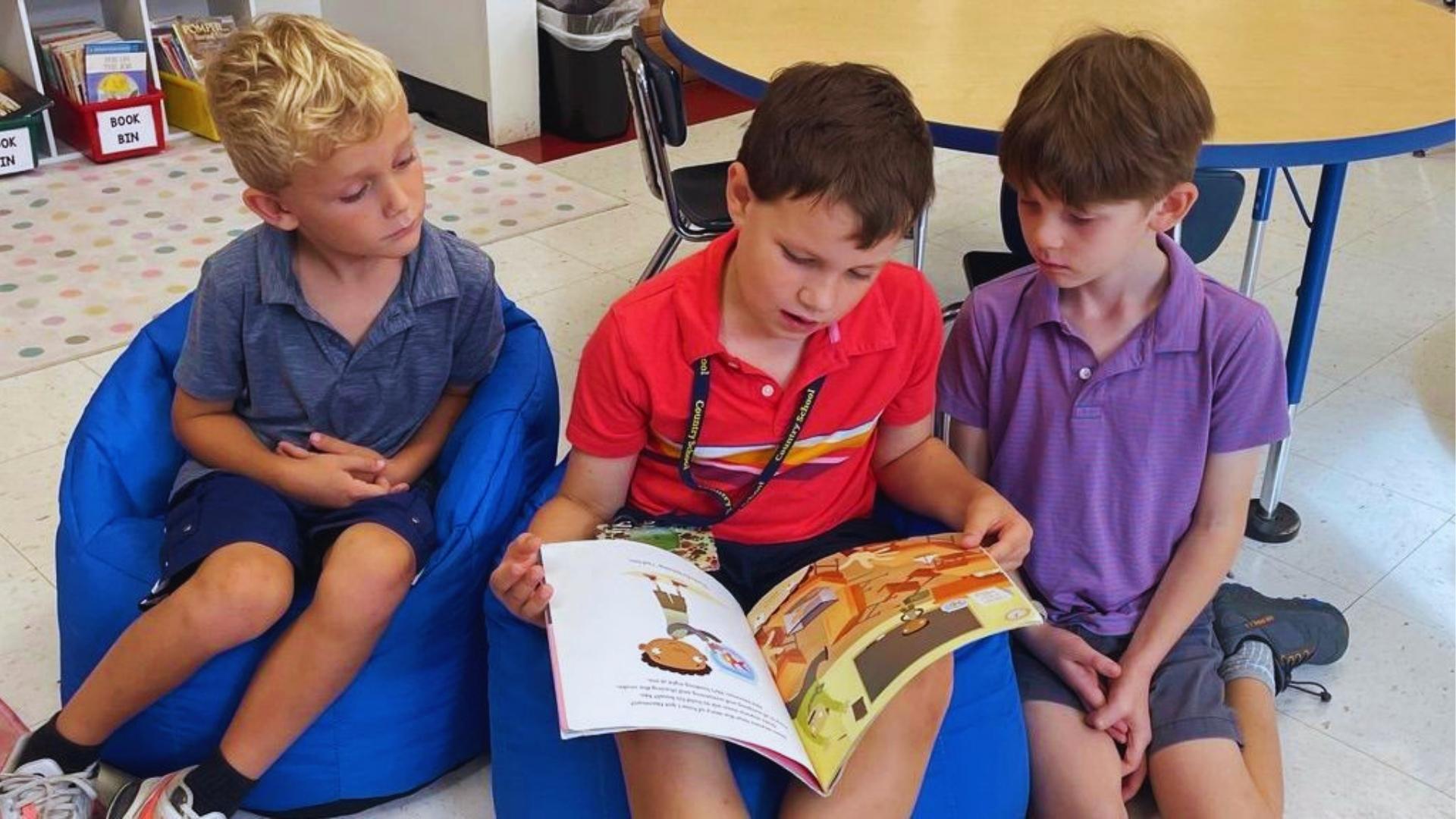 Three young boys in the classroom sitting on blue beanbags sharing colorful picture books during reading time.