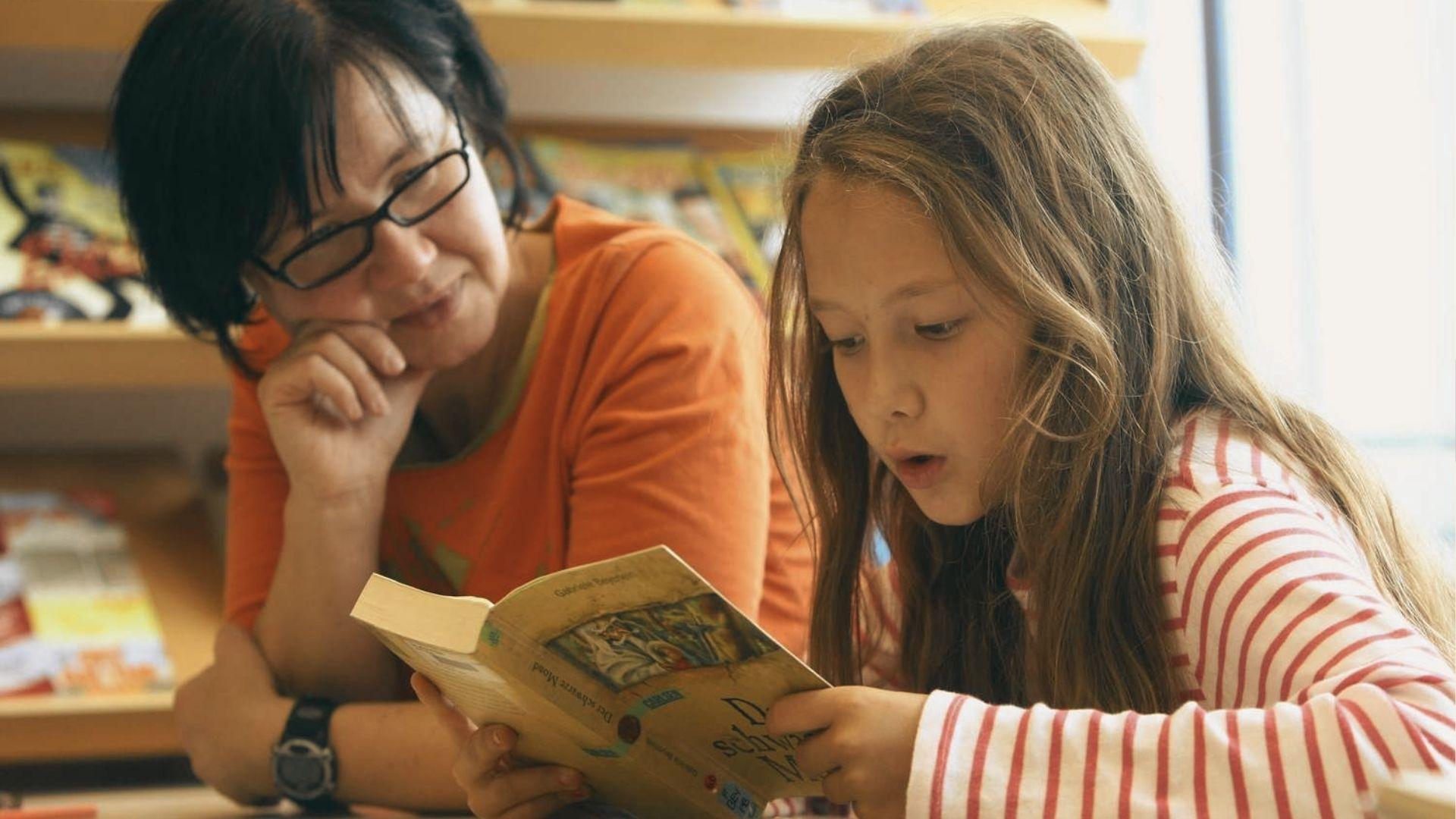 Teacher reads aloud from a book while a young girl listens intently in a library.