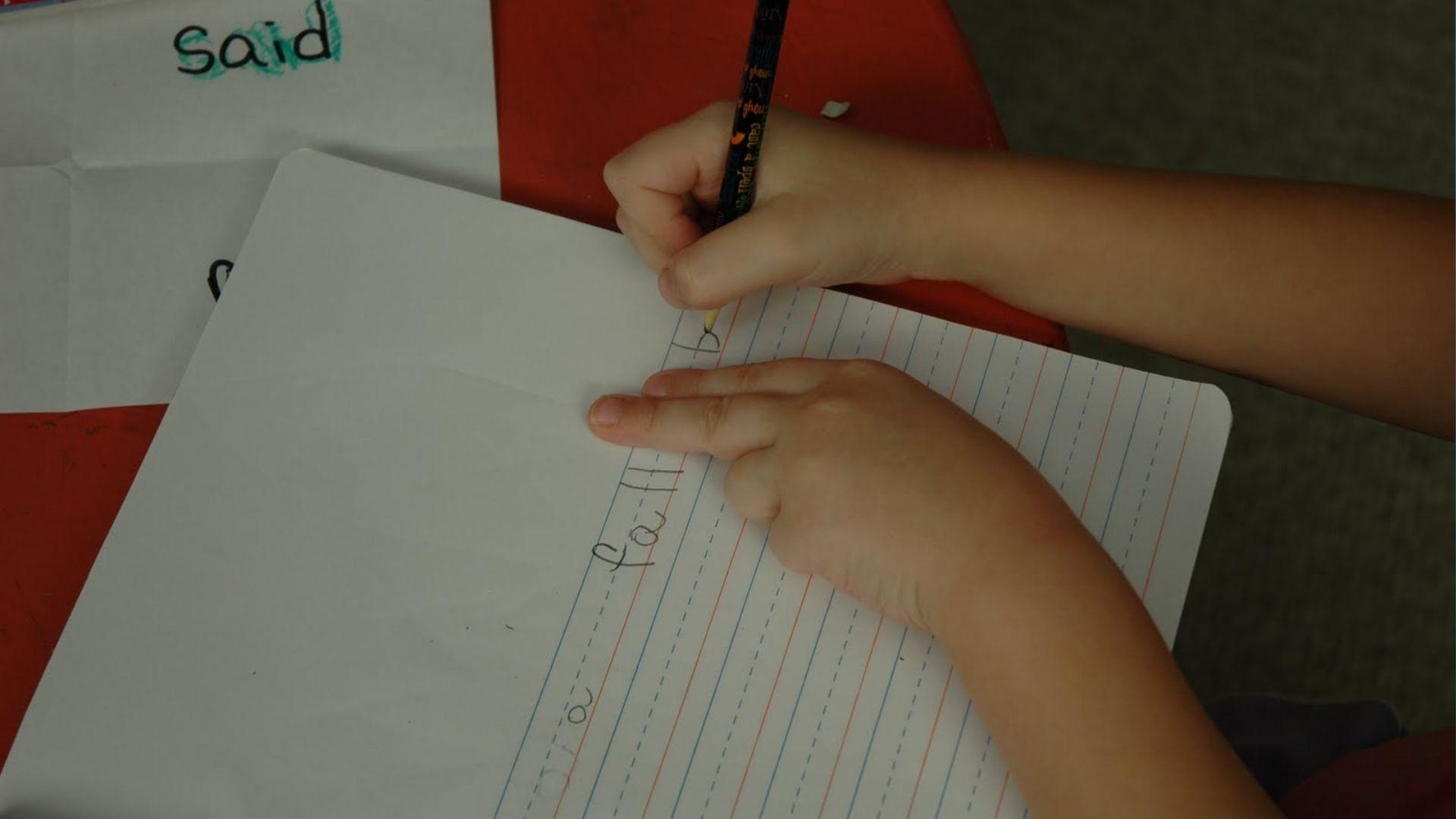 Child’s hands write letters on ruled paper with finger spacing guide, teaching letter alignment.