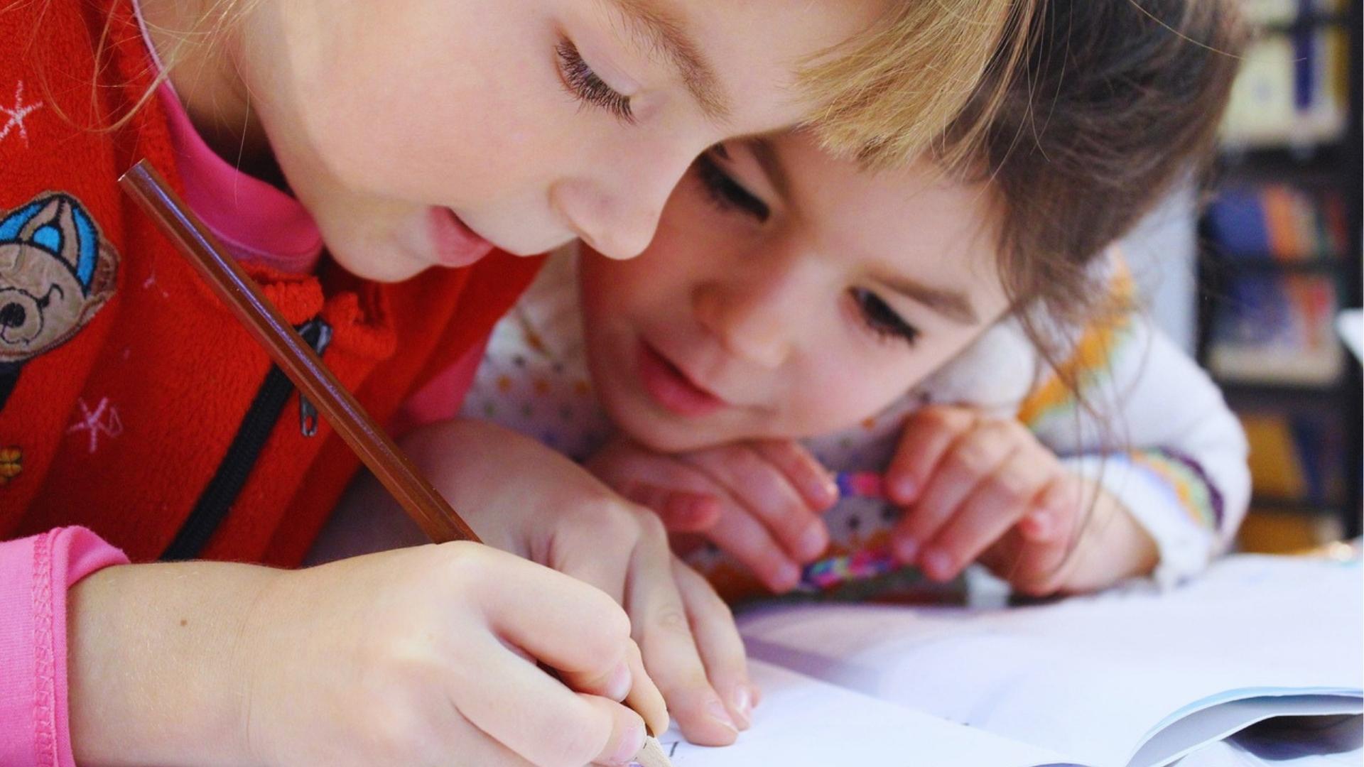 Two young girls sharing a desk, one writing with brown pencil while the other watches and smiles.
