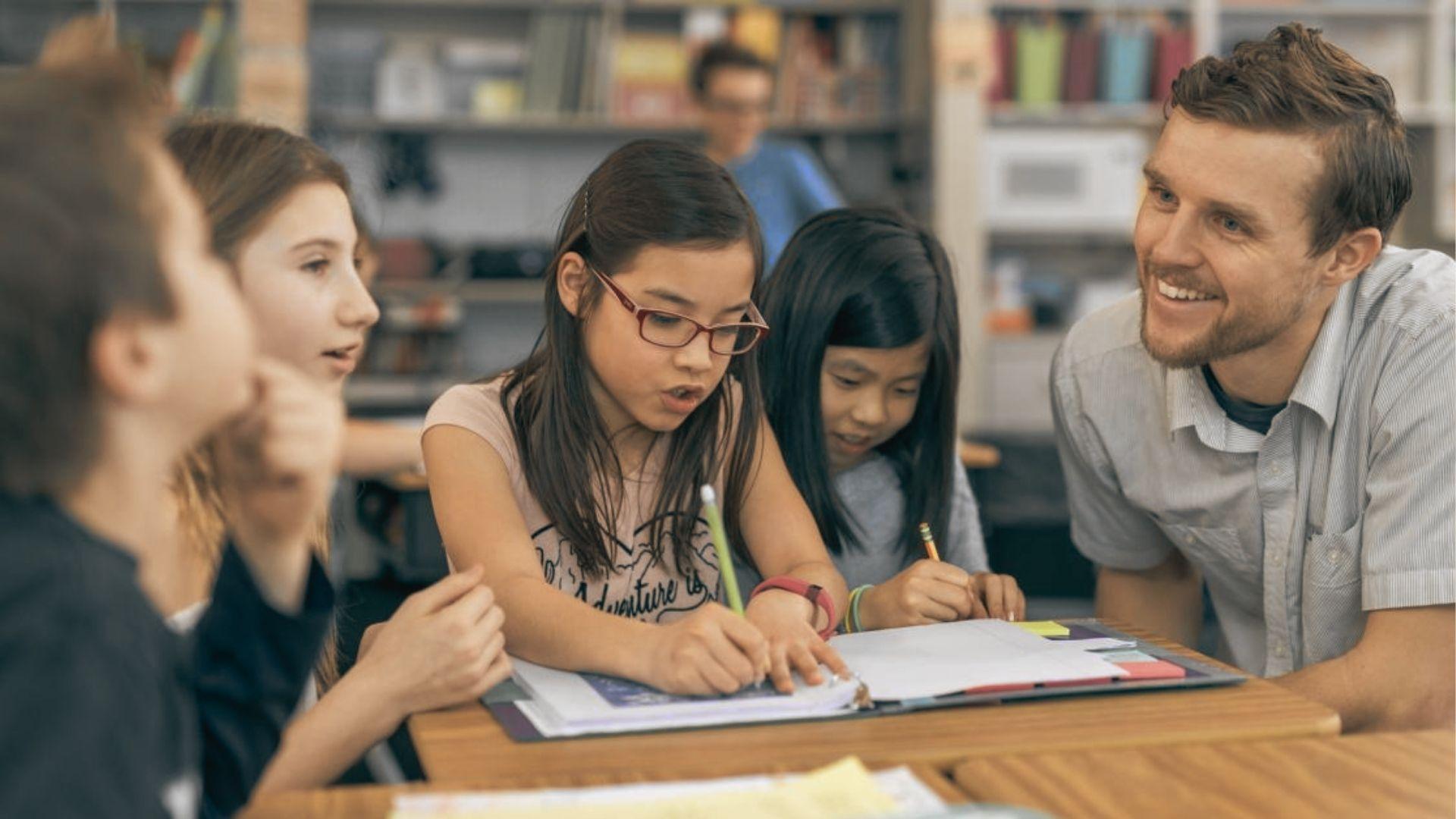 Teacher kneels beside a small group of students gathered around a book in a library setting.