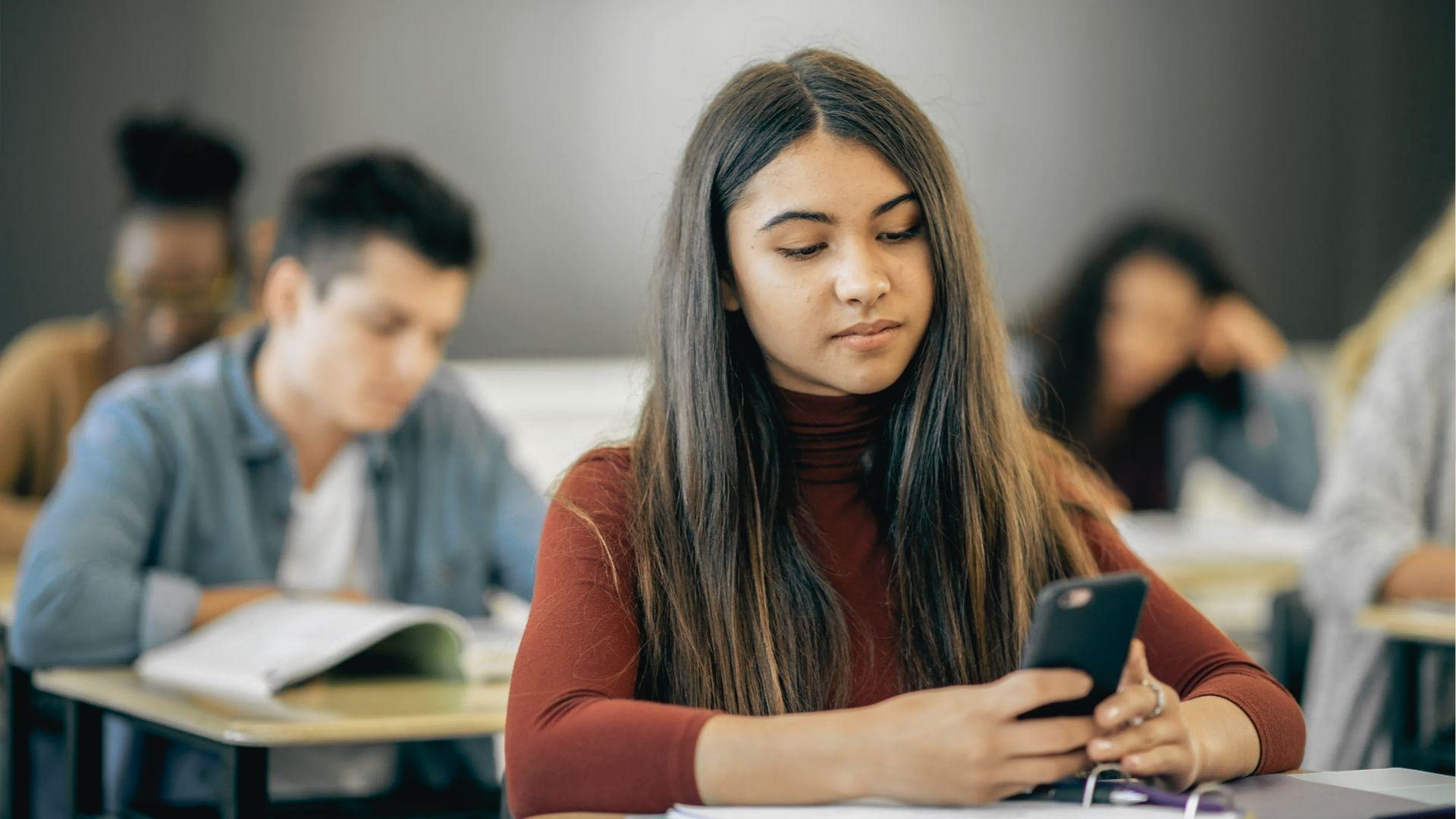 Teenage girl in a red turtleneck looks at her phone while sitting in a classroom.