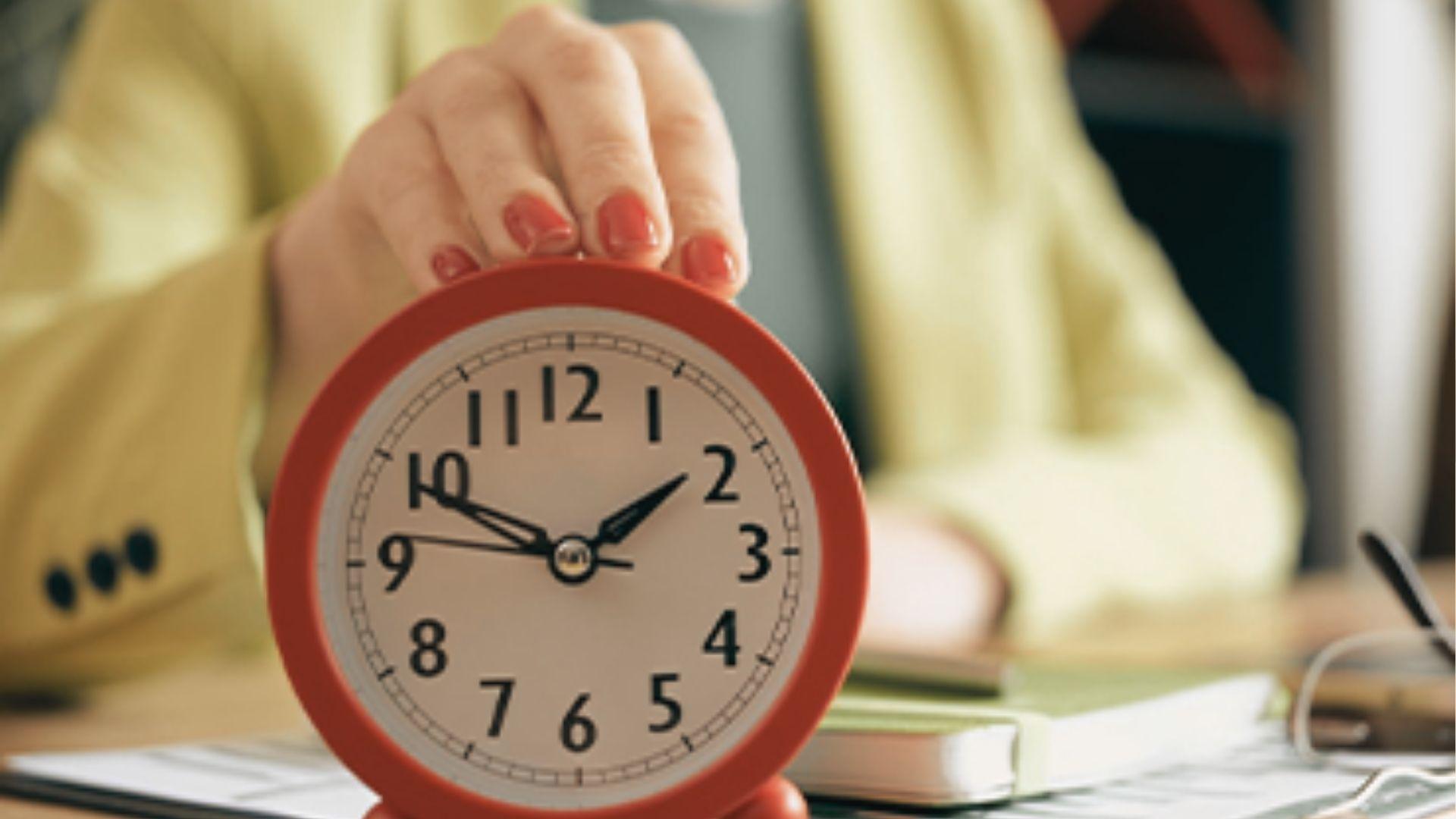 Hand with red nail polish holds a red analog alarm clock above a desk with books.