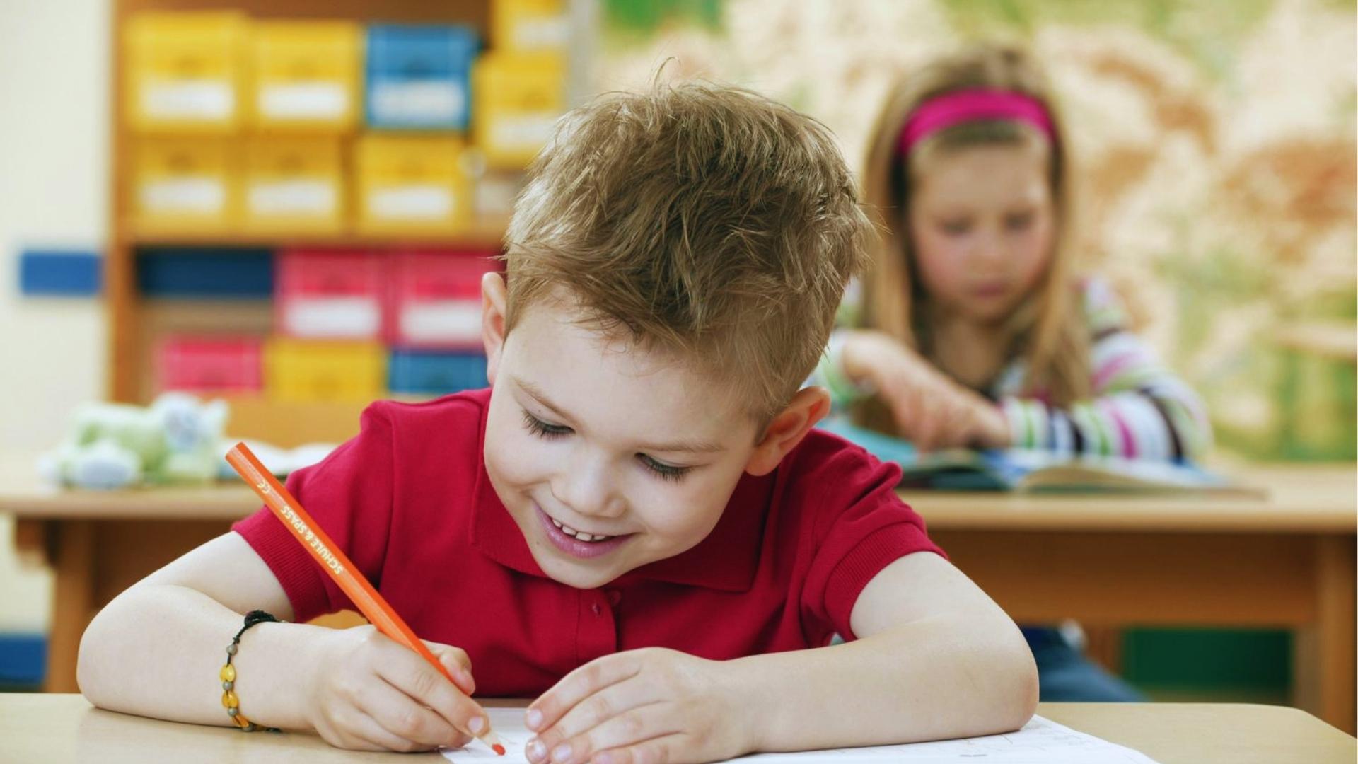 Smiling boy in red polo shirt writing with orange pencil at classroom desk with girl reading behind.