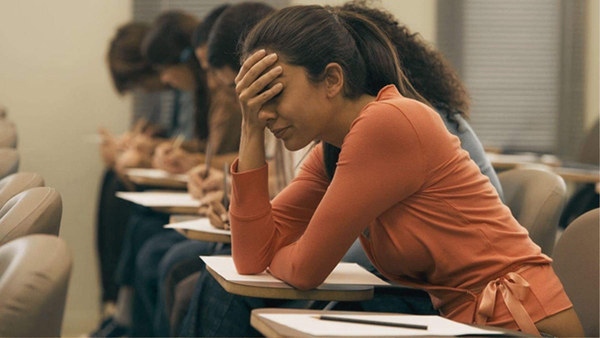 Stressed female student in an orange shirt holds her head during an exam with classmates behind her.